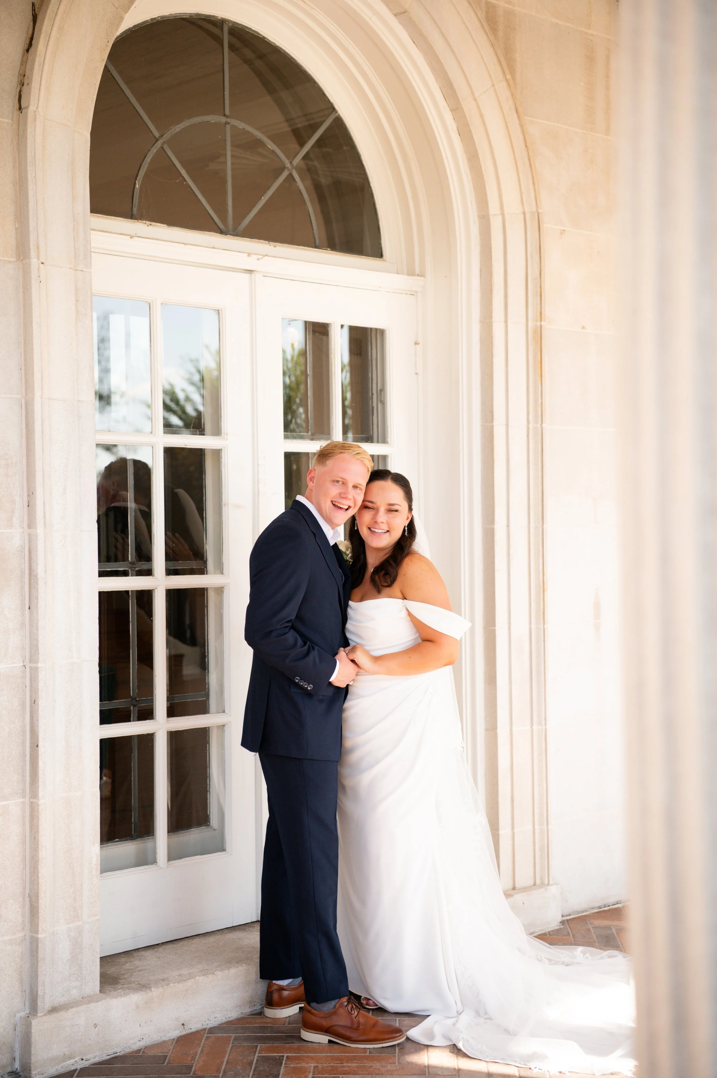 Bride in a stunning off the shoulder gown posing with groom against an elegant stone mansion.