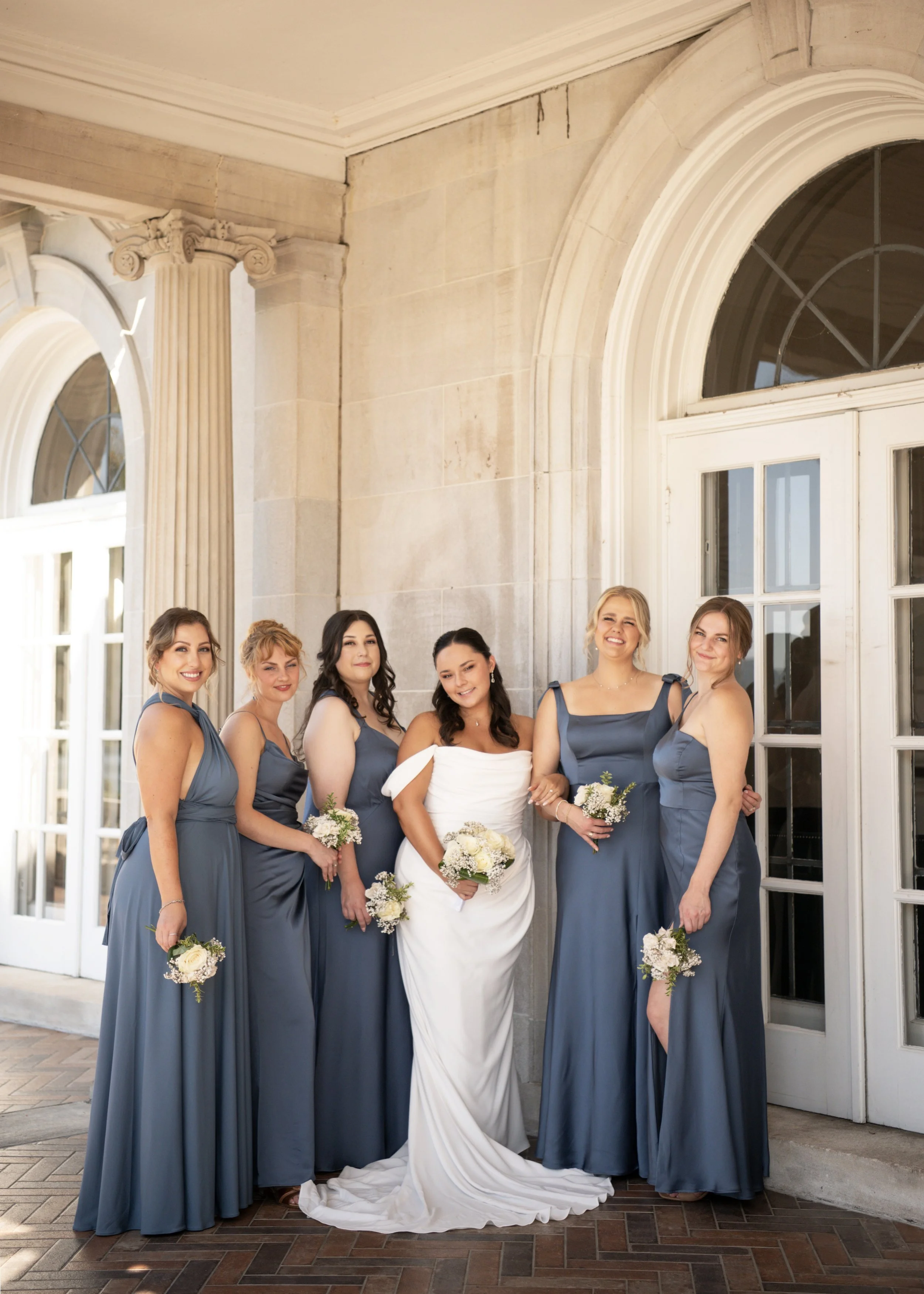 Bride with bridesmaids in dusty blue dresses posing outside of an elegant stone wedding structure.