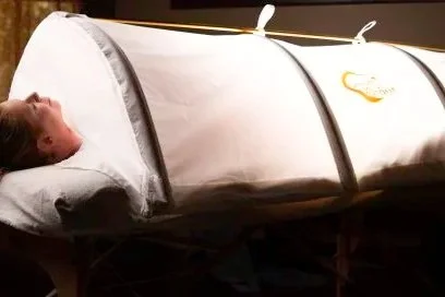 Person lying in a portable sleep pod or tent with a pillow in a dark room.