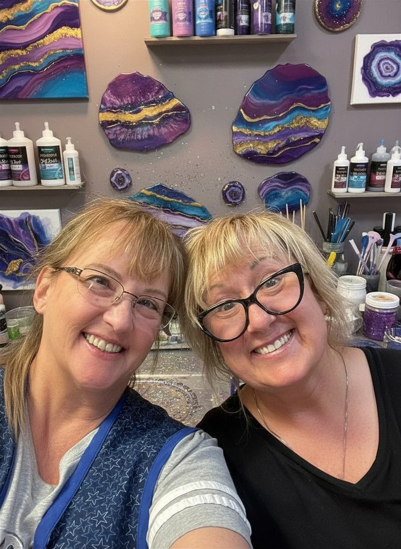 Two smiling women with glasses taking a selfie in front of a wall decorated with purple and gold geode-inspired art pieces and art supplies.