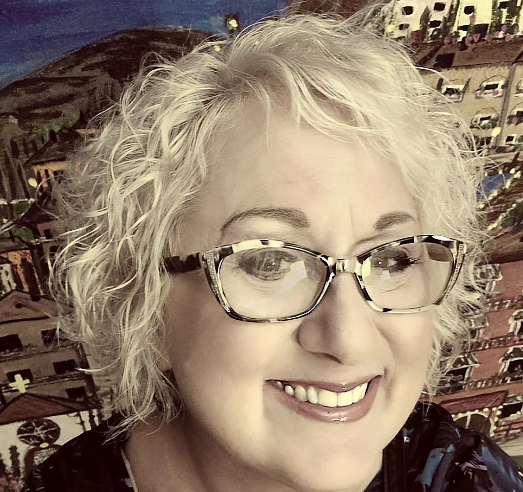 A woman with curly blonde hair and glasses smiling in front of a colorful, artistic mural of a cityscape.