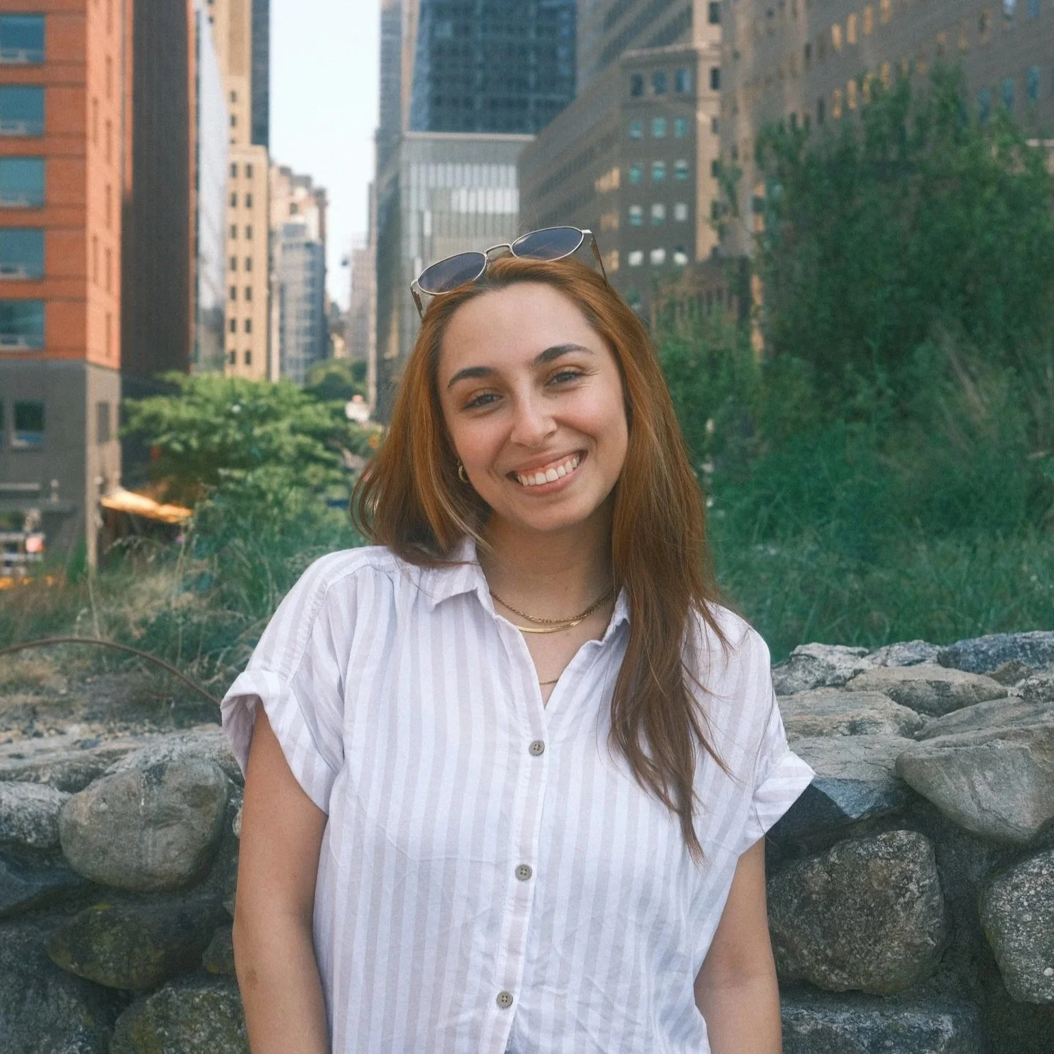 Headshot of Valeria, blog writer of Everything About Coffee. She has read hair, sunglasses on her head. A white vertically striped shirt, with a city background.