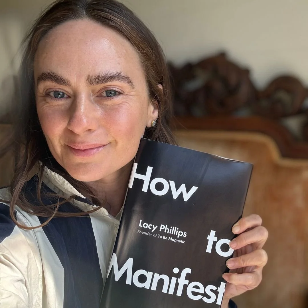 A woman holding a book titled 'How to Manifest' by Lacy Phillips, smiling at the camera.