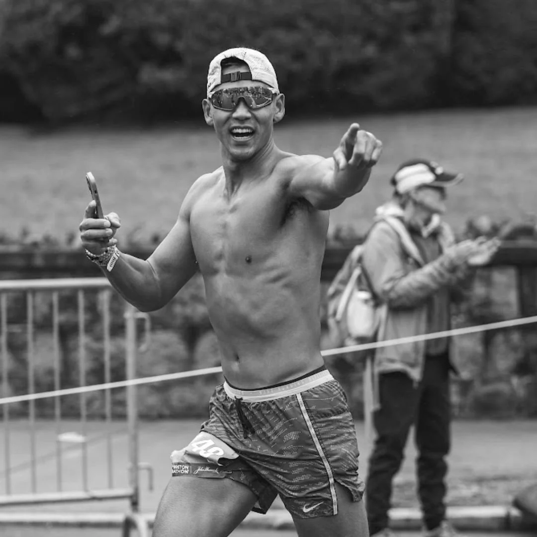 A shirtless male runner in athletic shorts and sunglasses gestures with his right hand during a race, with a woman in the background using her phone.