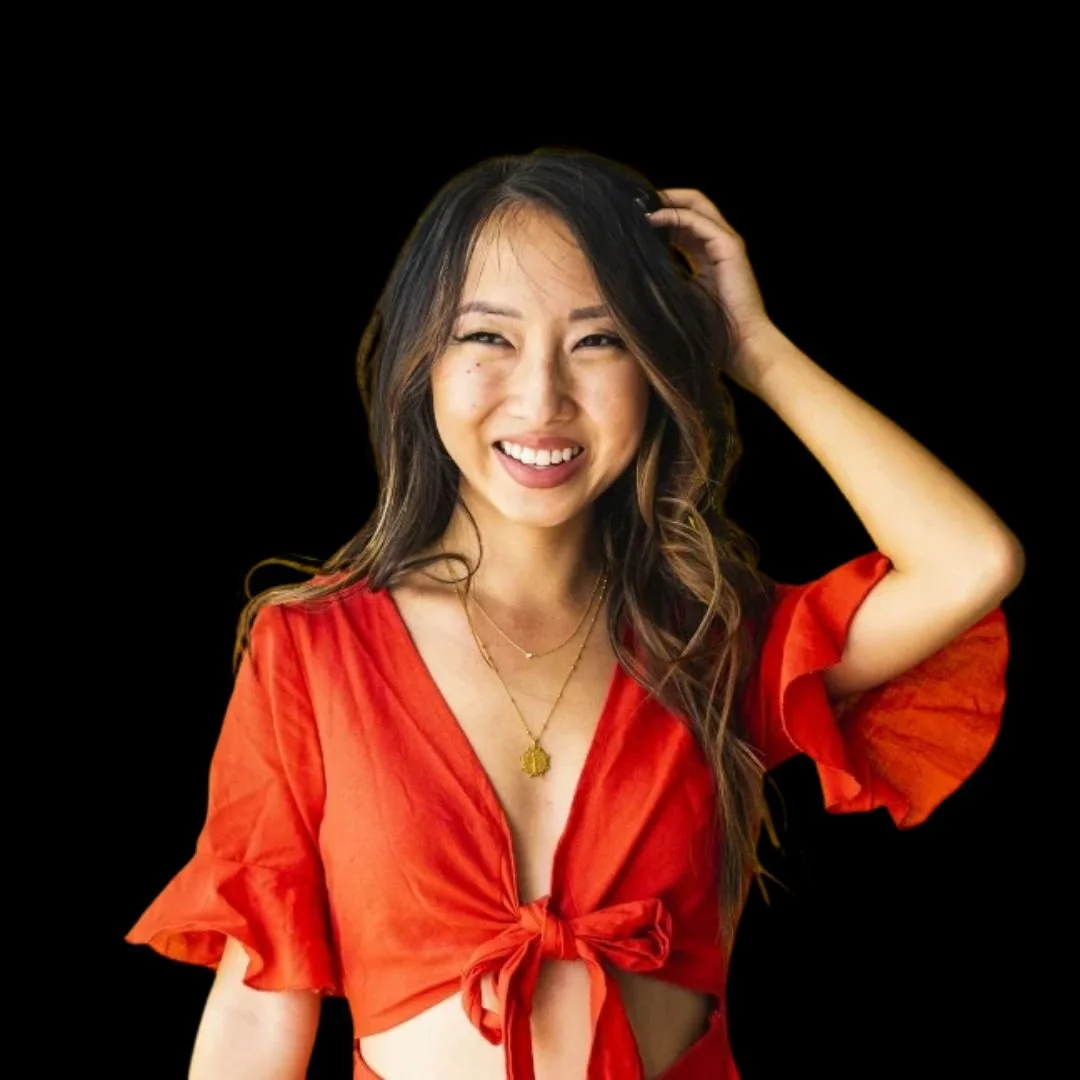 Young woman with long, wavy hair wearing a red tied top, smiling and scratching her head against a black background.