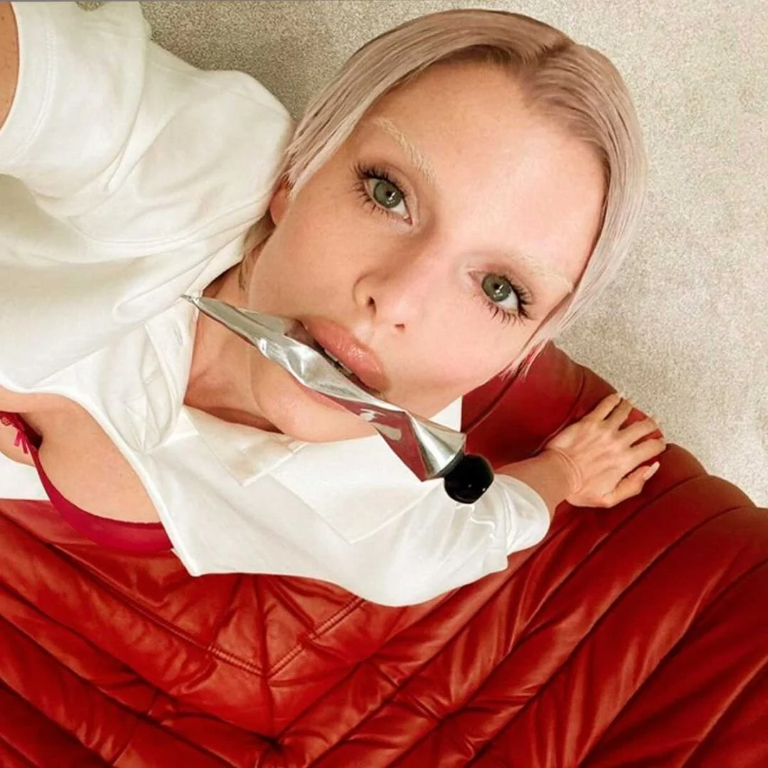 A woman with blonde hair, green eyes, wearing a white shirt and red leather pants, holding a large knife in her mouth, sitting on a red leather sofa.