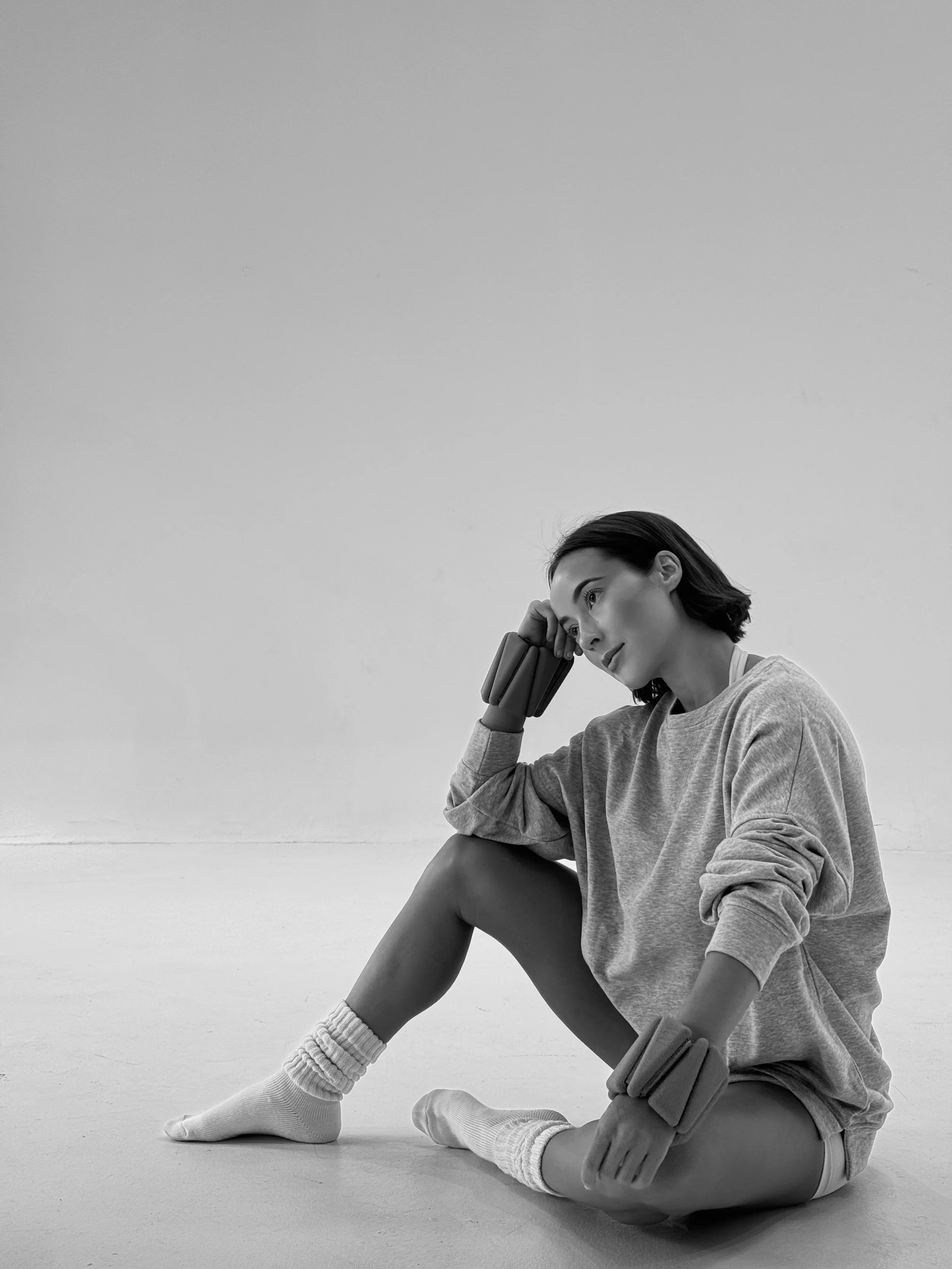 A black and white photo of a young woman sitting on the floor in a stretching pose, with her right elbow resting on her knee and her hand supporting her head, wearing a loose sweatshirt, shorts, and knee-high socks.