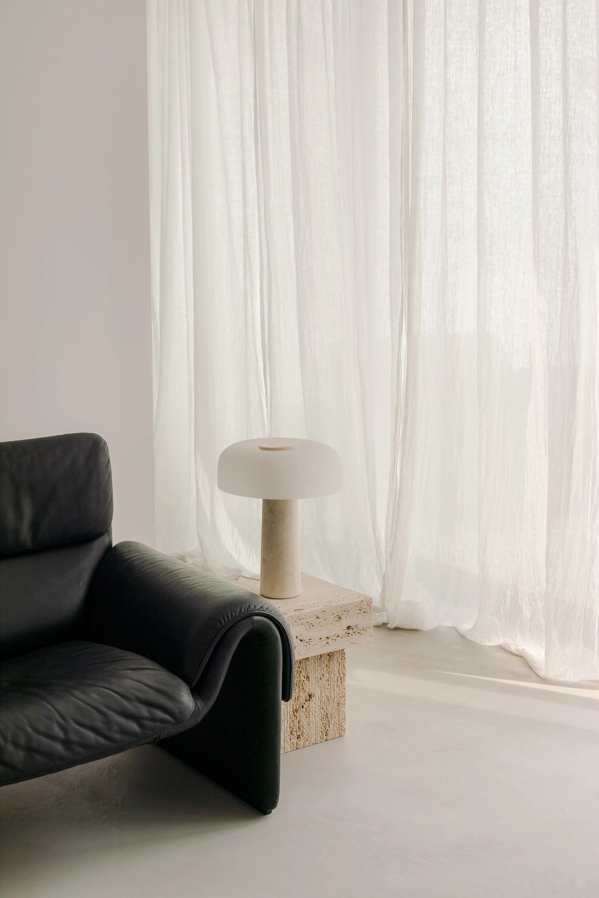 Living room corner with a black leather sofa, a stone side table, a lamp with a marble base, and sheer white curtains letting in natural light.