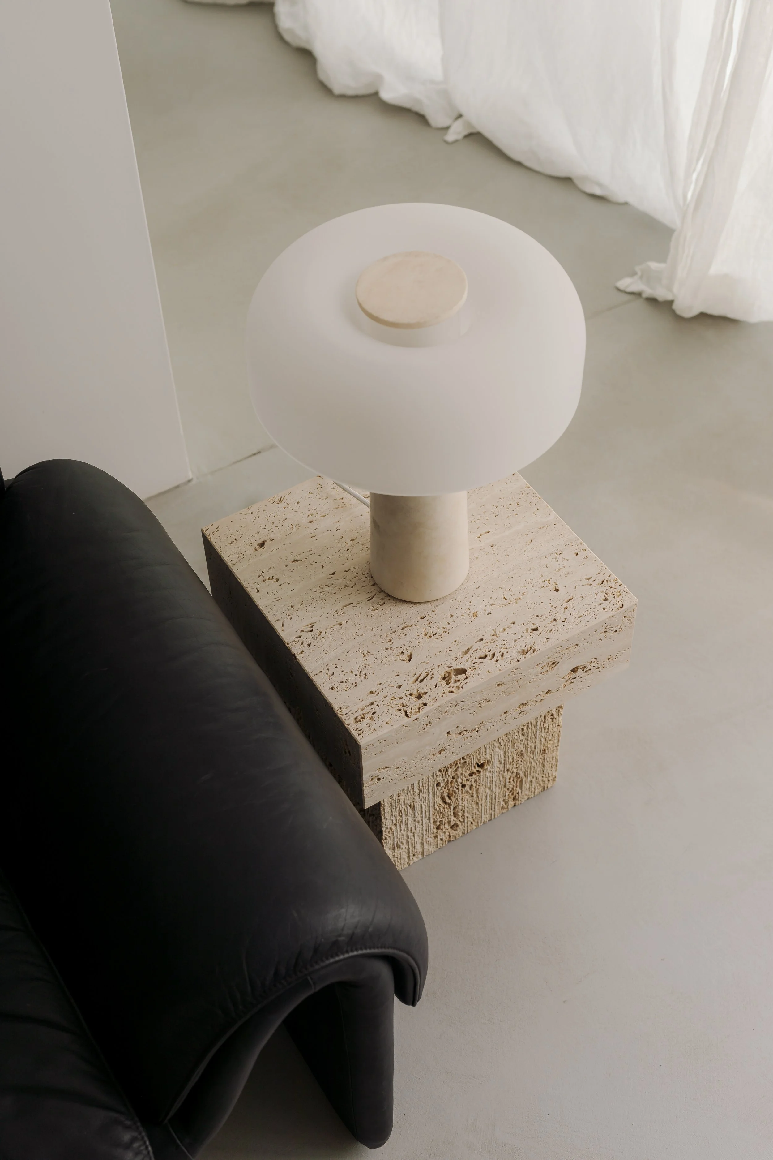 A small round table with a white tabletop and a beige base, placed on a textured beige pedestal, positioned next to a black leather armchair on a light-colored floor with a white curtain in the background.