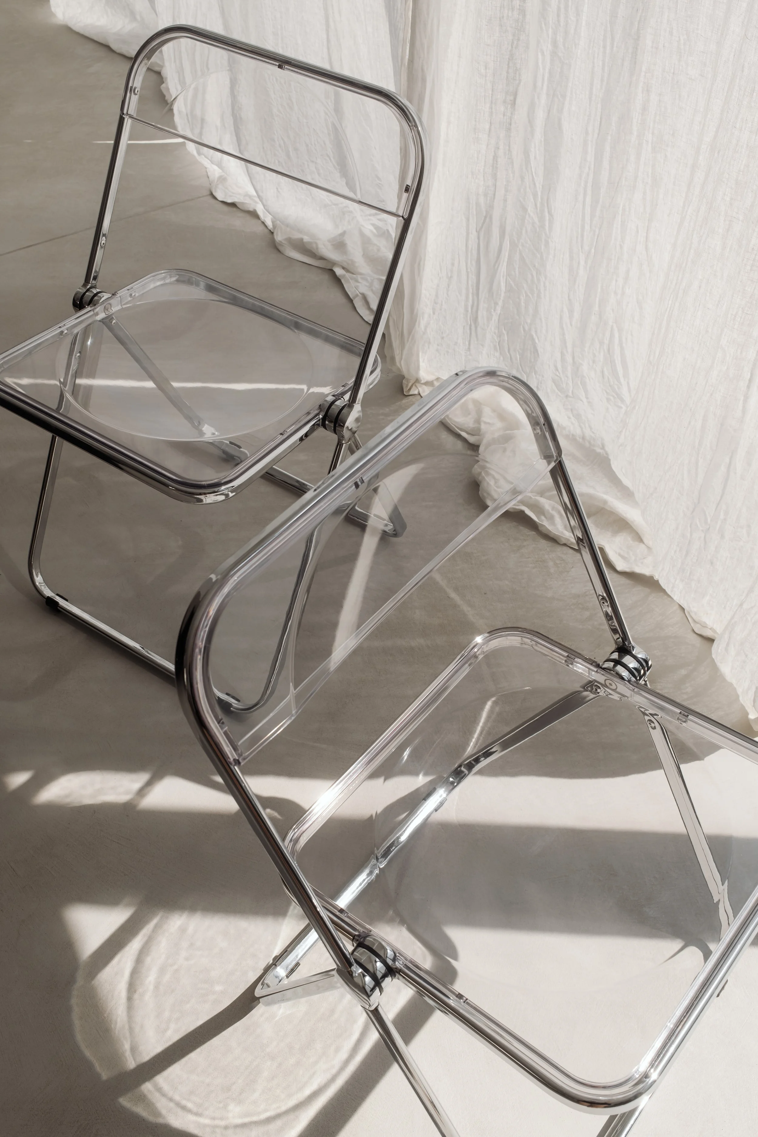 Two transparent folding chairs with metal frames arranged on a light-colored concrete floor near a white fabric backdrop.