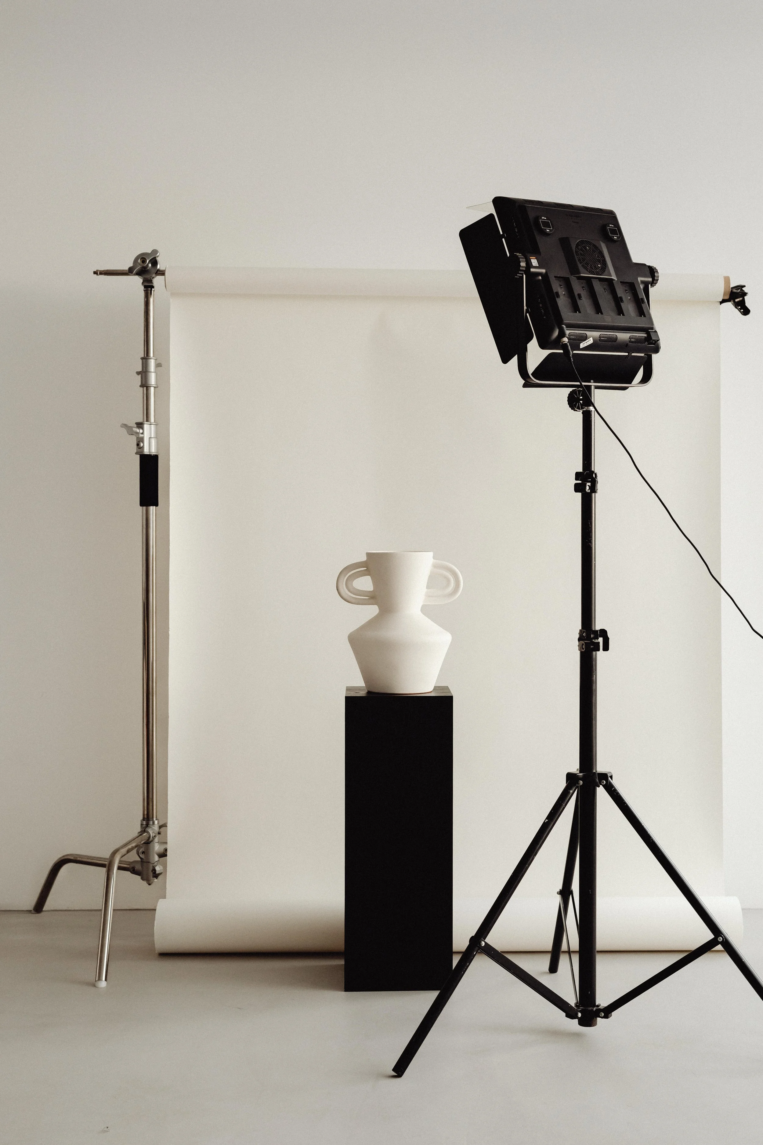 Photography studio setup with a black softbox light on a stand, a white ceramic vase with handles on a black pedestal, a silver and black backdrop stand, all against a white background.