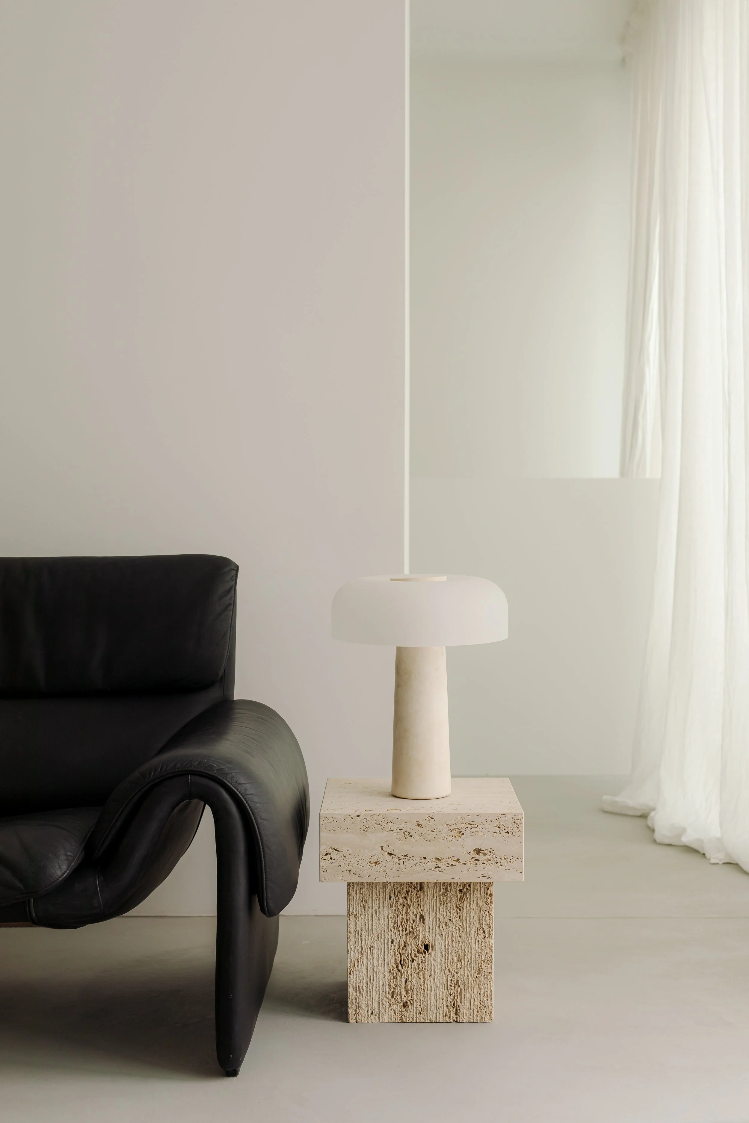 Interior room with a black leather armchair on the left, a white table lamp on a stone pedestal in the center, and sheer white curtains on the right.