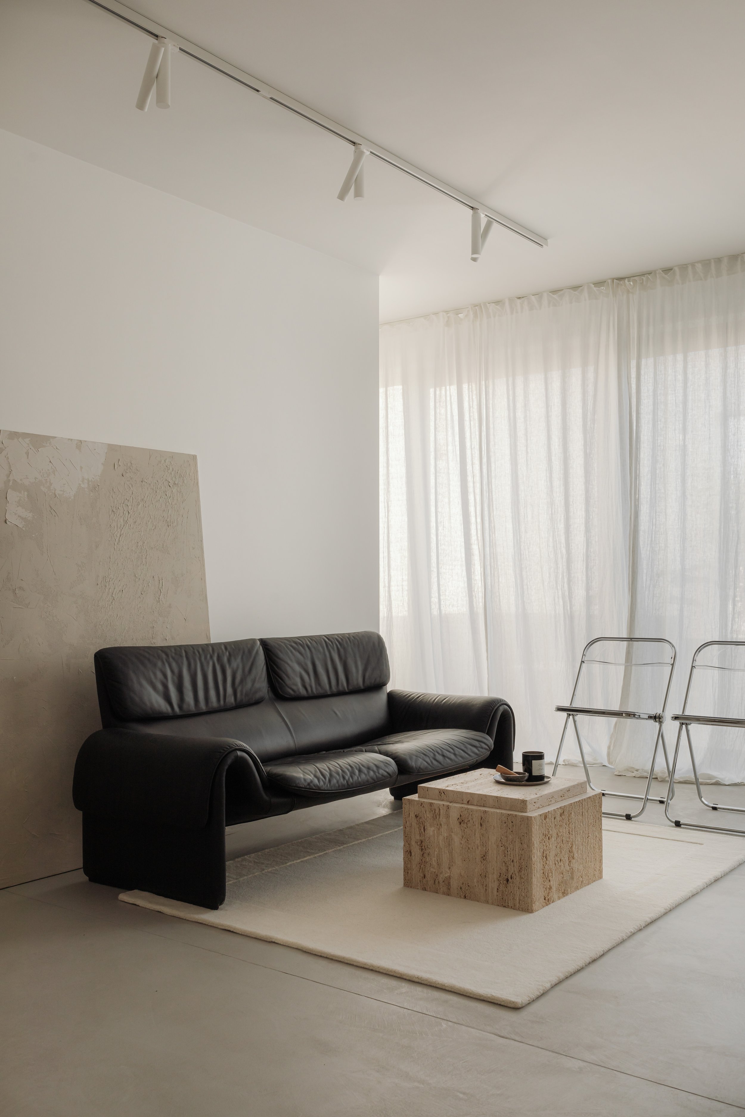 A modern, minimalist living room with a black leather sofa, a textured abstract wall art piece, a rectangular stone coffee table, and two transparent acrylic chairs near a large window with white curtains, illuminated by natural light.