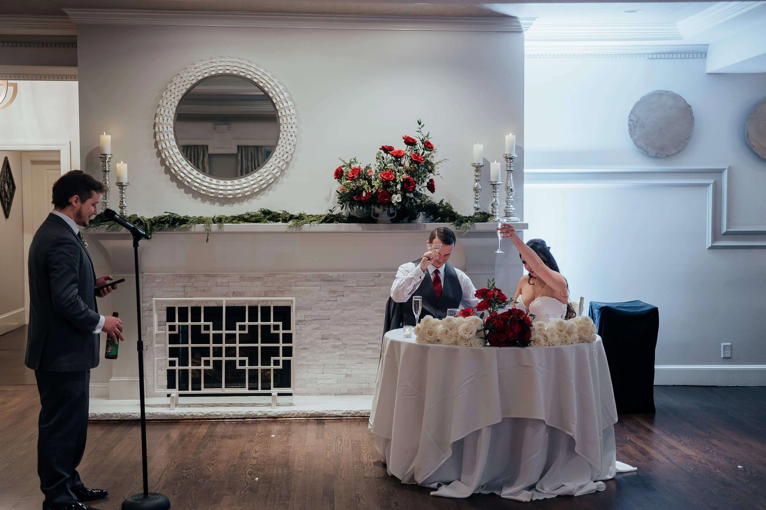 Bride and groom toasting at their sweetheart table adorned with red and white rose arrangements beside a greenery-draped fireplace at Saphire Estate, Sharon Massachusetts wedding florals by Milkweed Floral Co