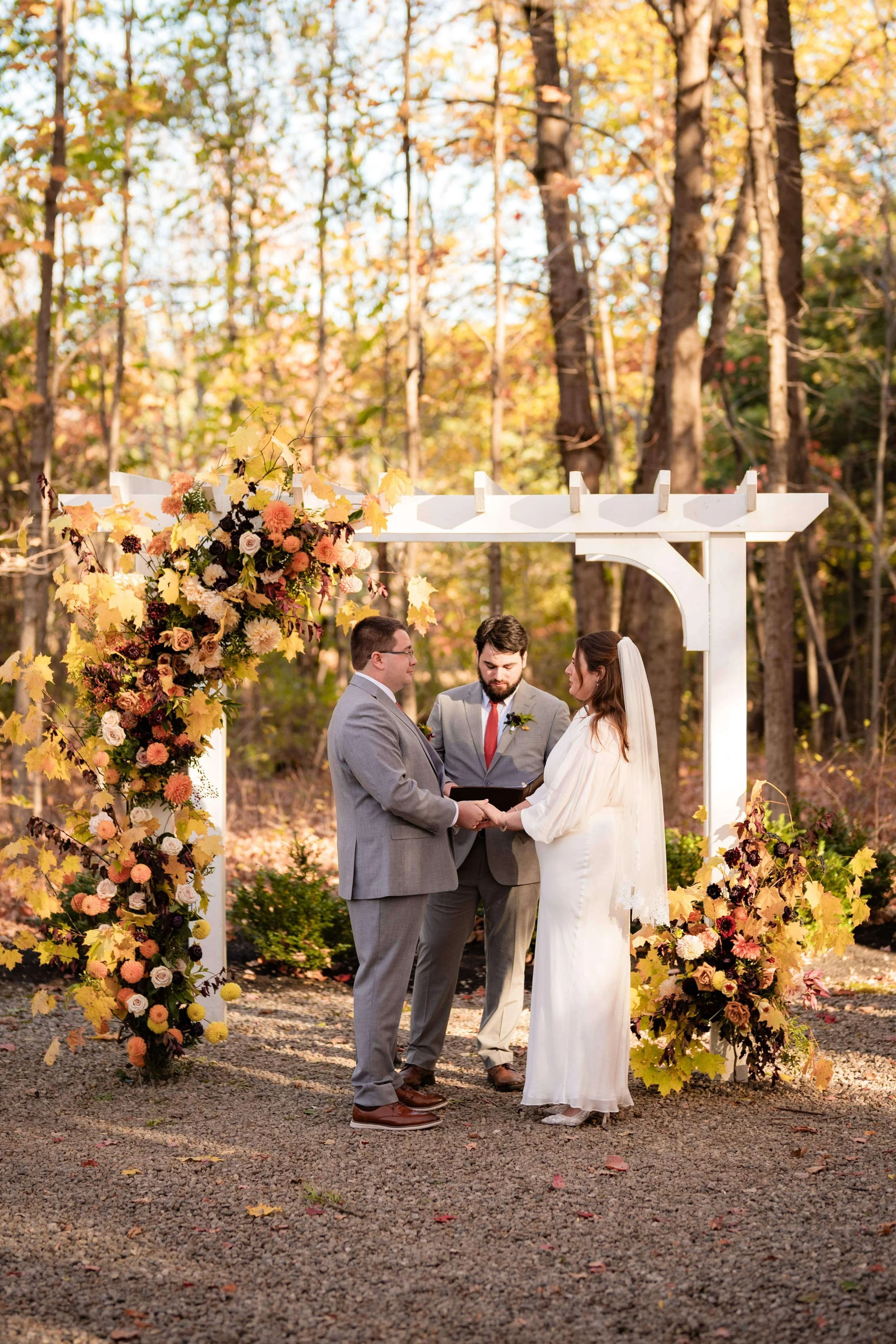 Bride and groom with officiant standing under white arbor decorated with yellow, peach, and orange dahlias at golden hour, fall wedding, Maine, florals by Milkweed Floral Co