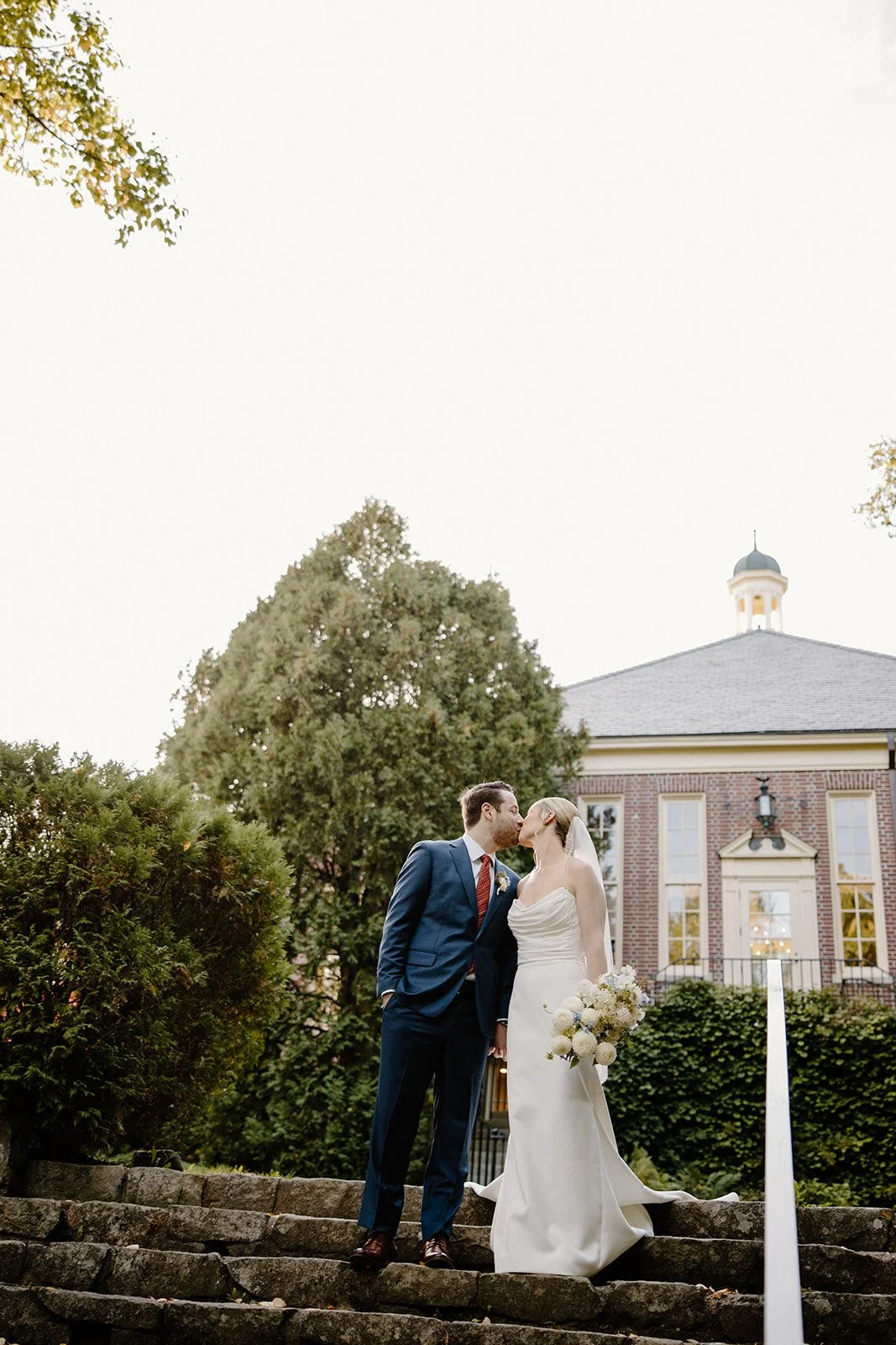 Bride and groom kissing on the stone steps of Camden Public Library, bride holding a white dahlia and blue delphinium bouquet, coastal Maine wedding by Milkweed Floral Co