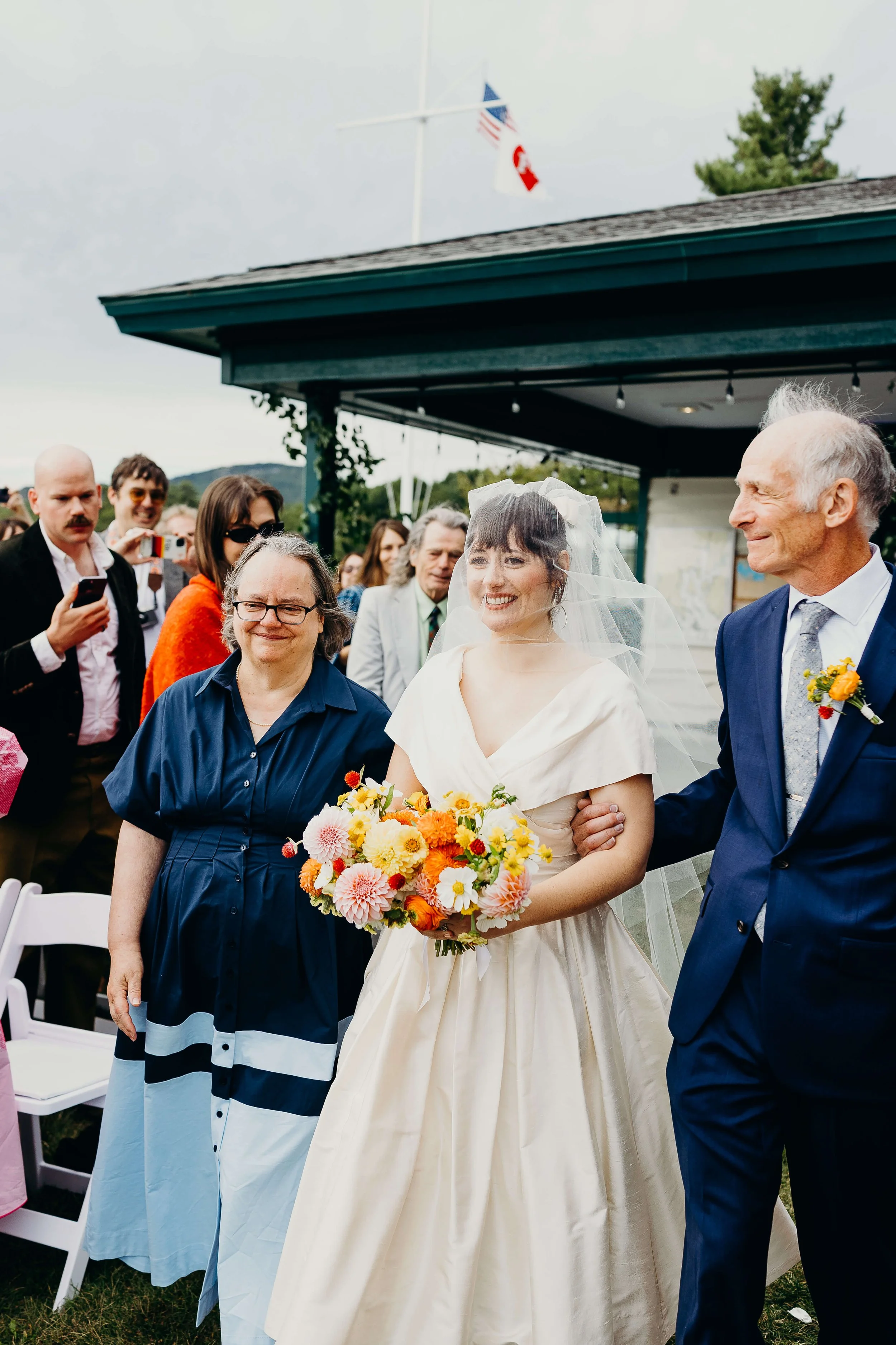 Bride walking down the aisle escorted by her parents, carrying a lush wildflower bouquet, wedding florals by Milkweed Floral Co