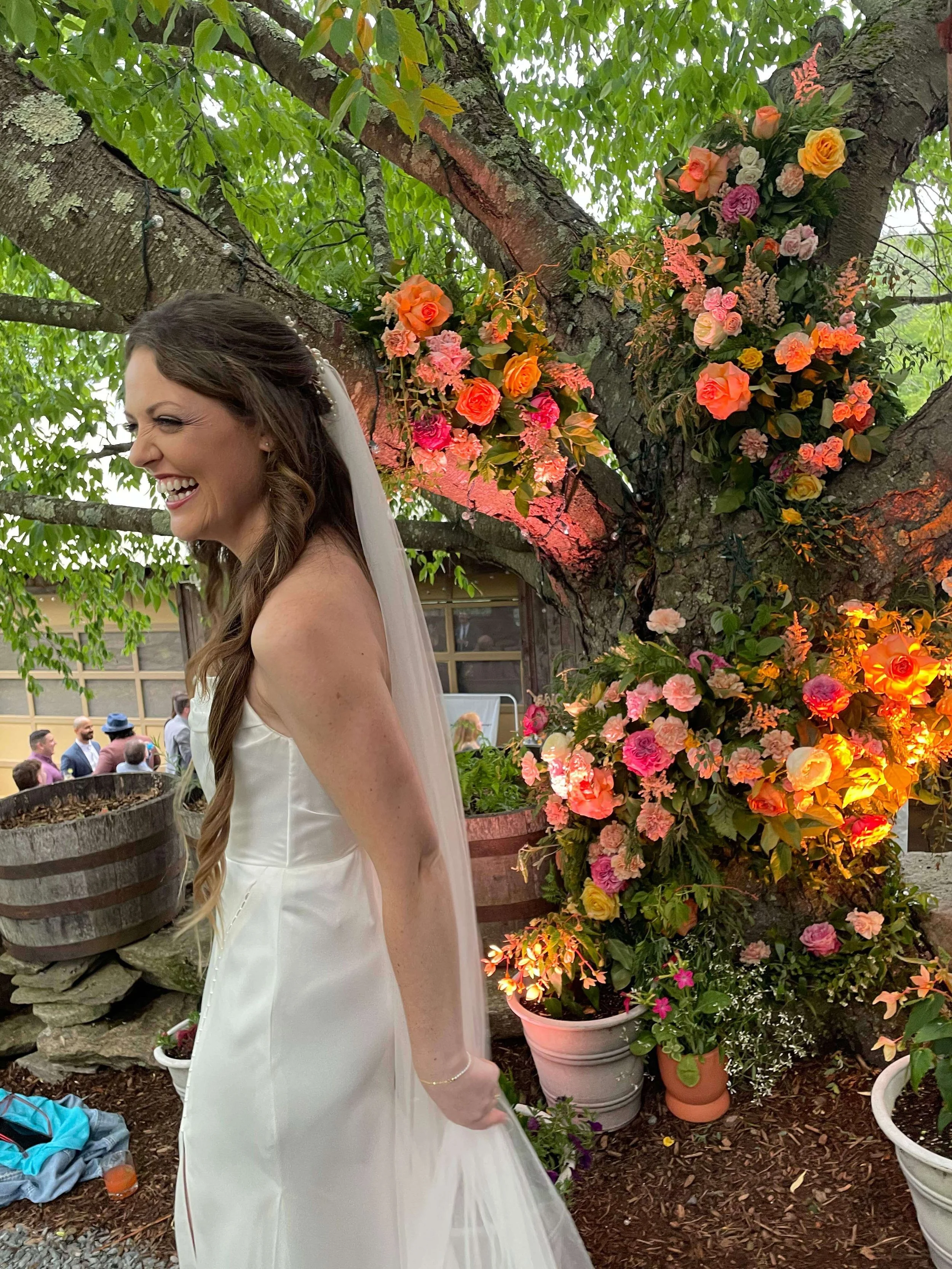 Laughing bride in front of a large floral installation of orange and pink garden roses and wildflowers woven through a tree, Montague Retreat Massachusetts, Milkweed Floral Co