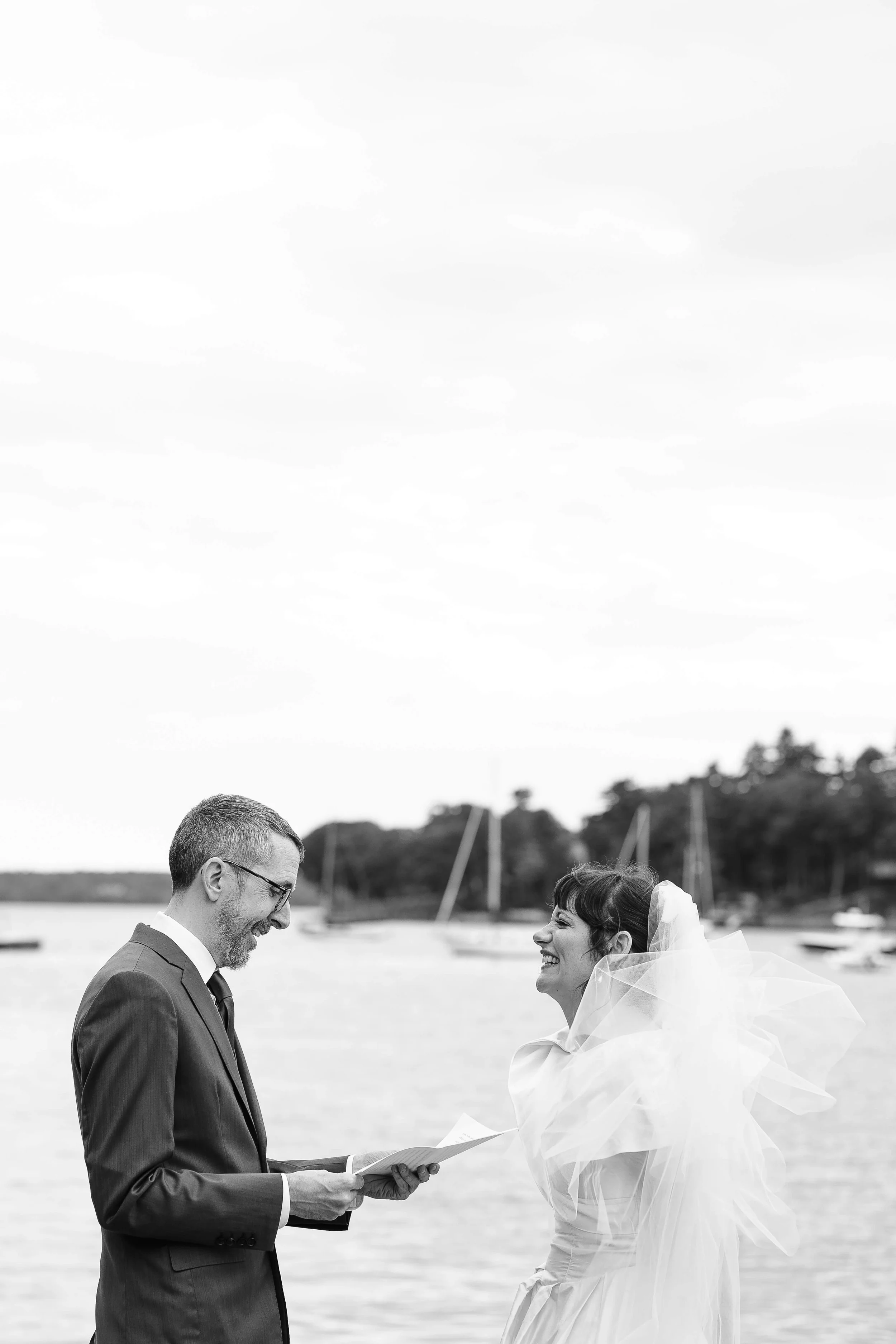 Groom reading vows as bride laughs with joy, waterfront ceremony with sailboats in the background in Blue Hill, Maine