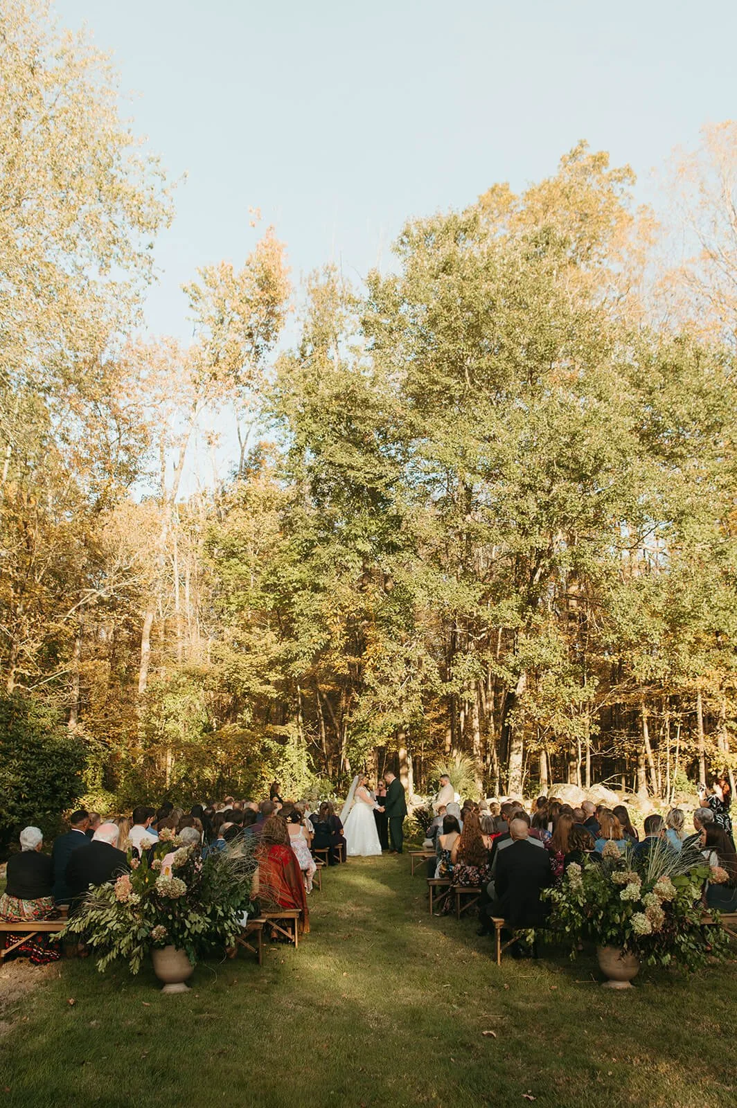 Wide view of an outdoor fall wedding ceremony with guests seated on wood benches flanked by large lush aisle arrangements of hydrangea, greenery, and ornamental grasses, Monson Massachusetts, Milkweed Floral Co