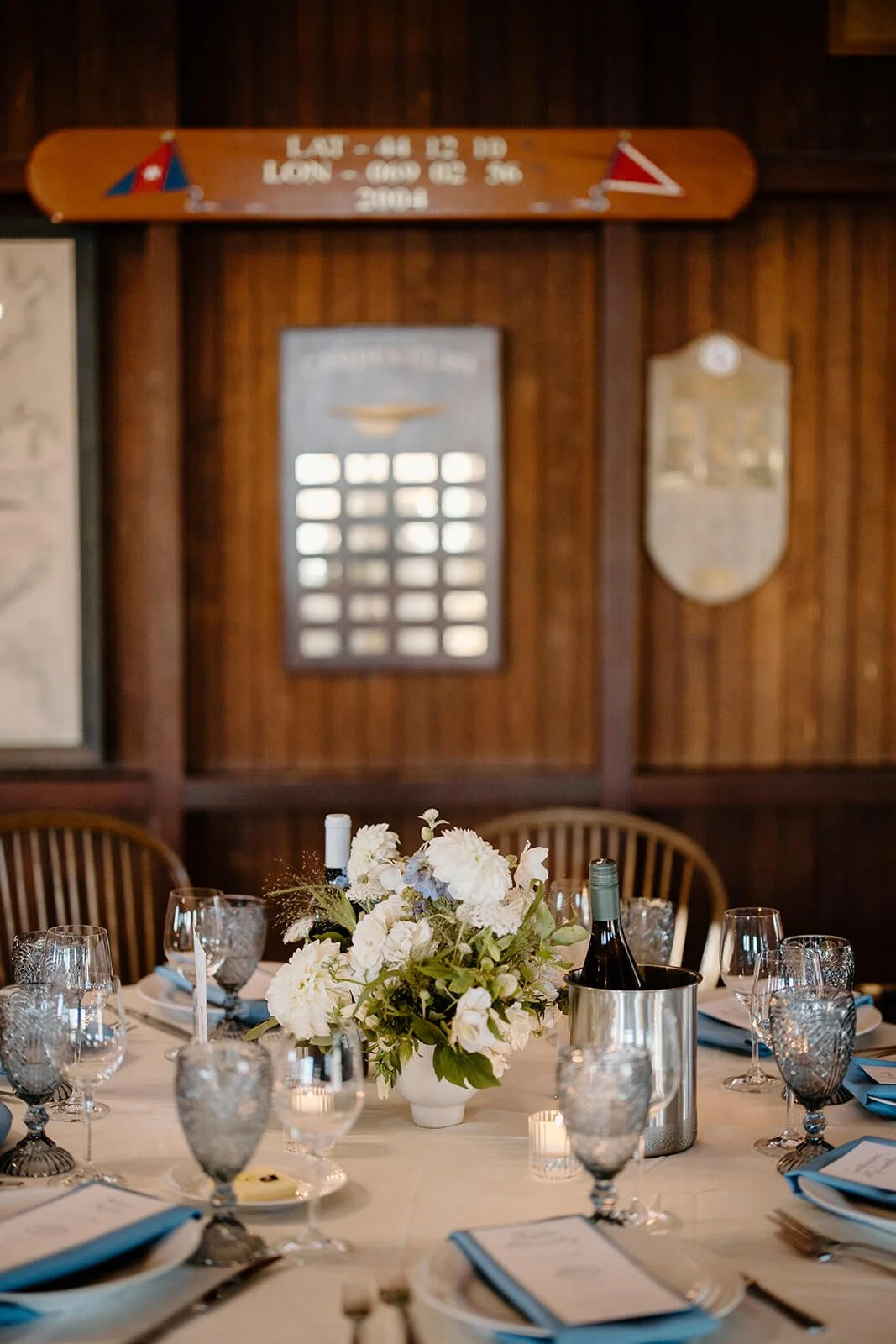 Classic coastal reception table at Camden Yacht Club with a white dahlia and hydrangea centerpiece, blue napkins, smoky glassware, and nautical decor on the walls, Milkweed Floral Co