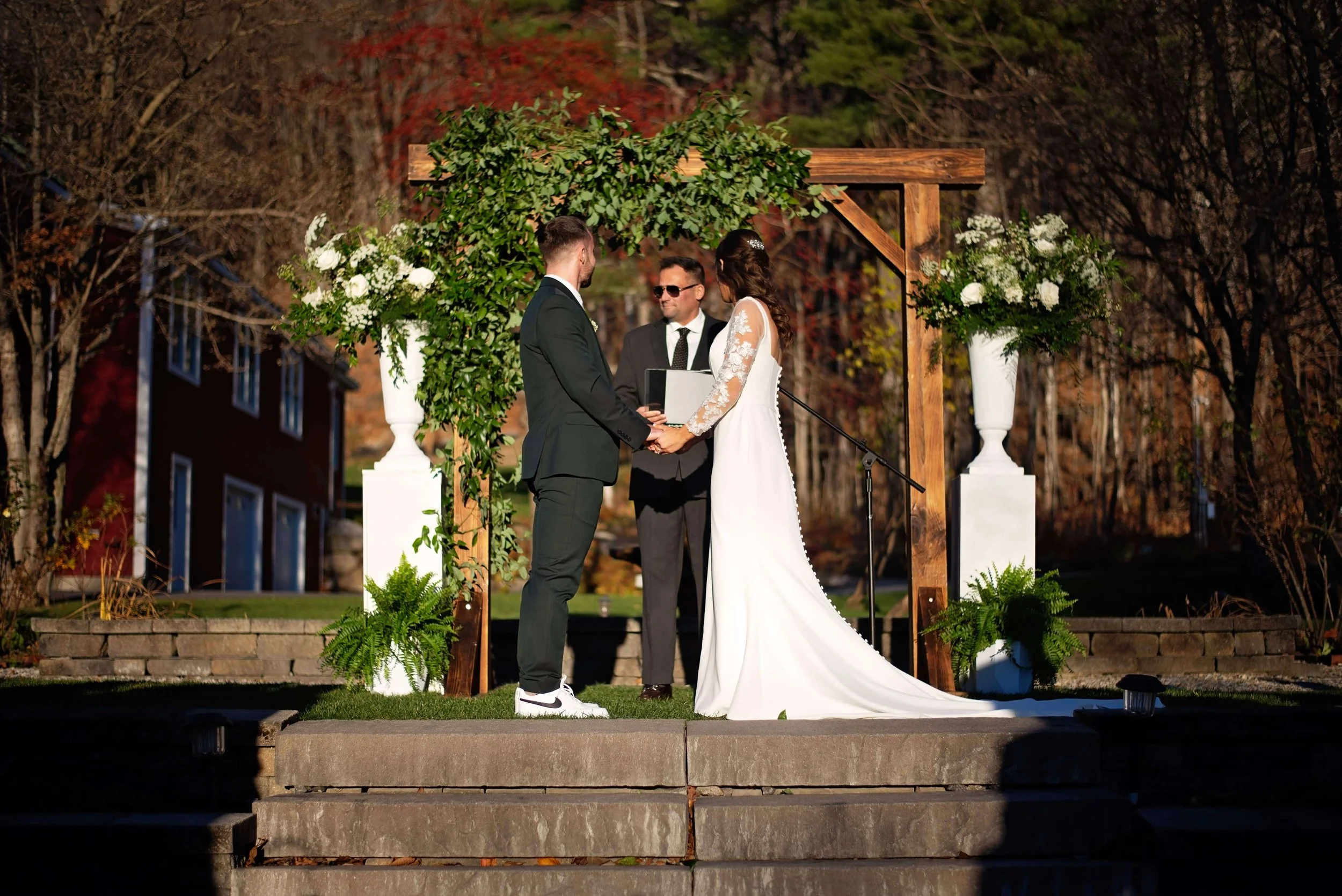 Bride and groom exchanging vows under a wood arbor draped with greenery and white floral arrangements at Newfound Lake Inn, Bridgewater New Hampshire wedding by Milkweed Floral Co