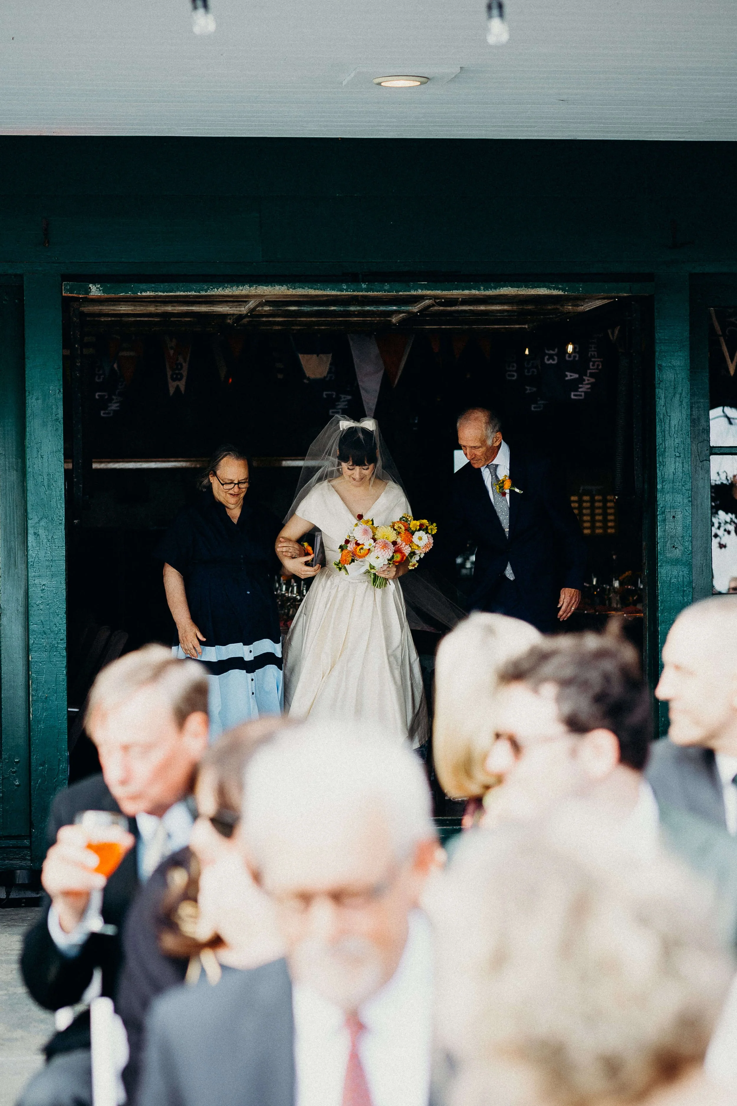 Bride walking out of Kollegewidgwok Yacht Club with her parents holding a vibrant orange, pink, and yellow wildflower bouquet, Blue Hill Maine wedding by Milkweed Floral Co