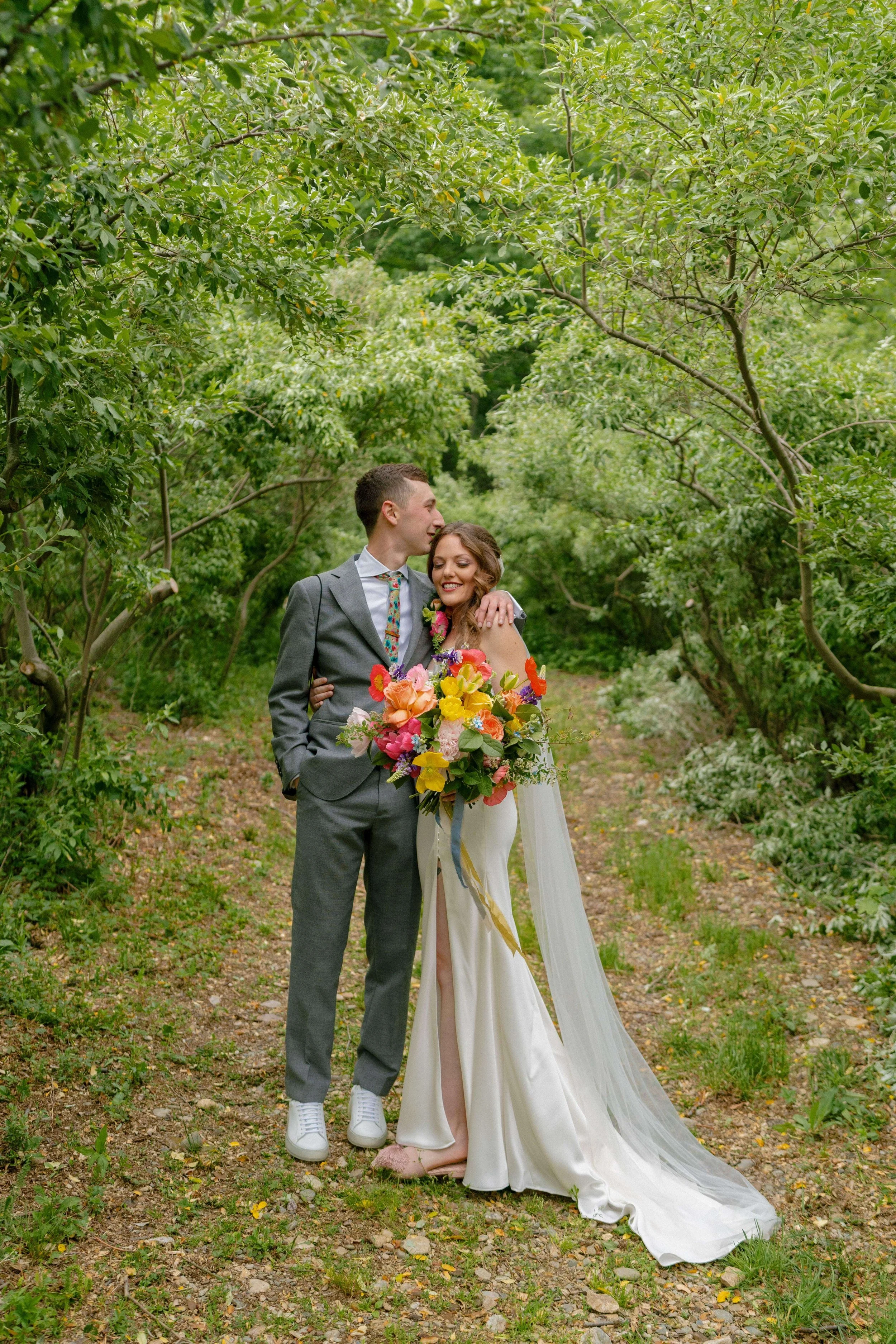Bride and groom embracing on a wooded path, bride holding a vibrant bouquet of orange poppies, yellow tulips, and pink wildflowers with ribbon streamers, Montague Massachusetts, Milkweed Floral Co