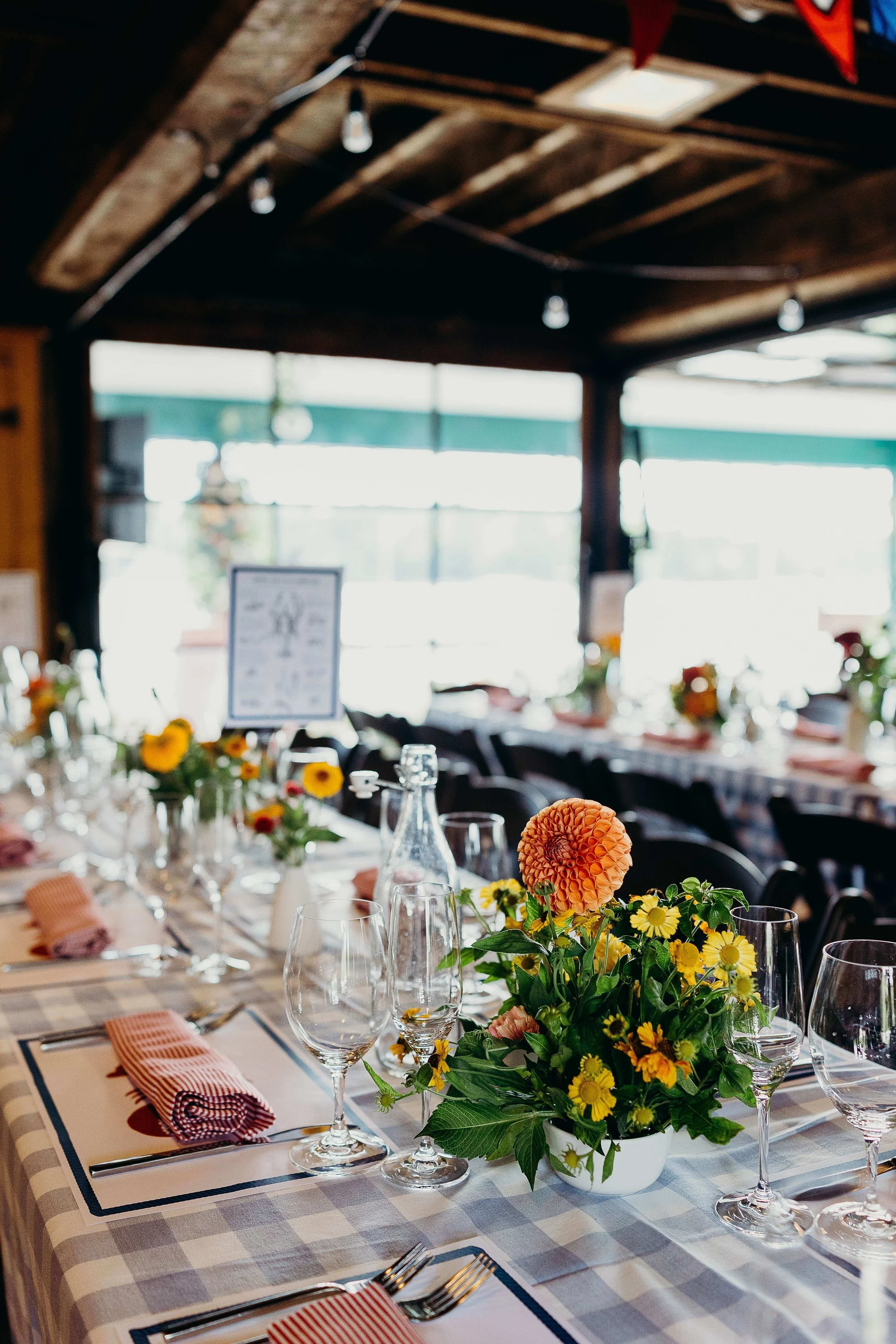 Long reception tables set with orange dahlia and yellow wildflower centerpieces, gingham tablecloths, and striped napkins at Kollegewidgwok Yacht Club, Blue Hill Maine wedding by Milkweed Floral Co
