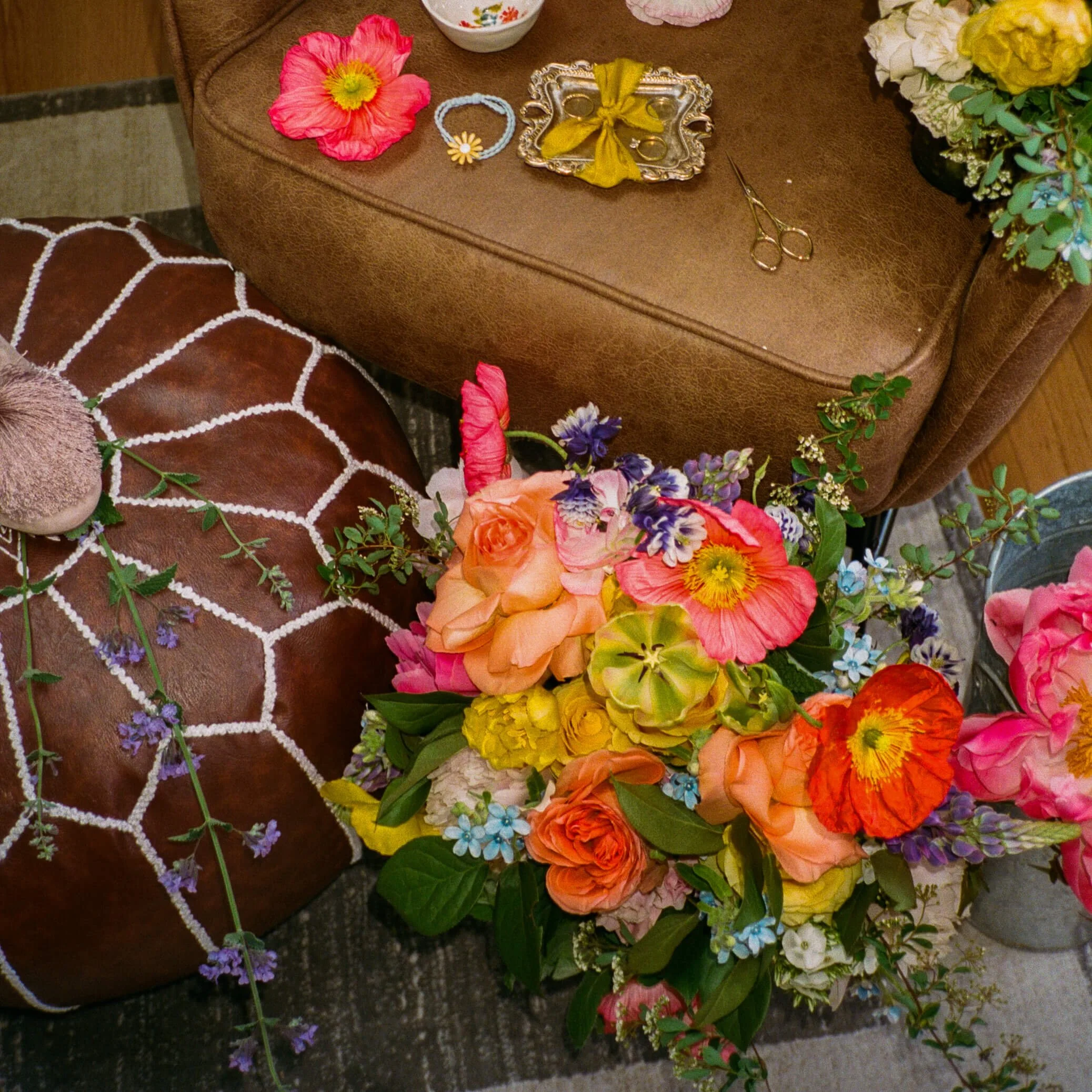 Vibrant wildflower bridal bouquet with pink poppies, orange garden roses, yellow tulips, and blue delphinium resting on a leather pouf, Milkweed Floral Co