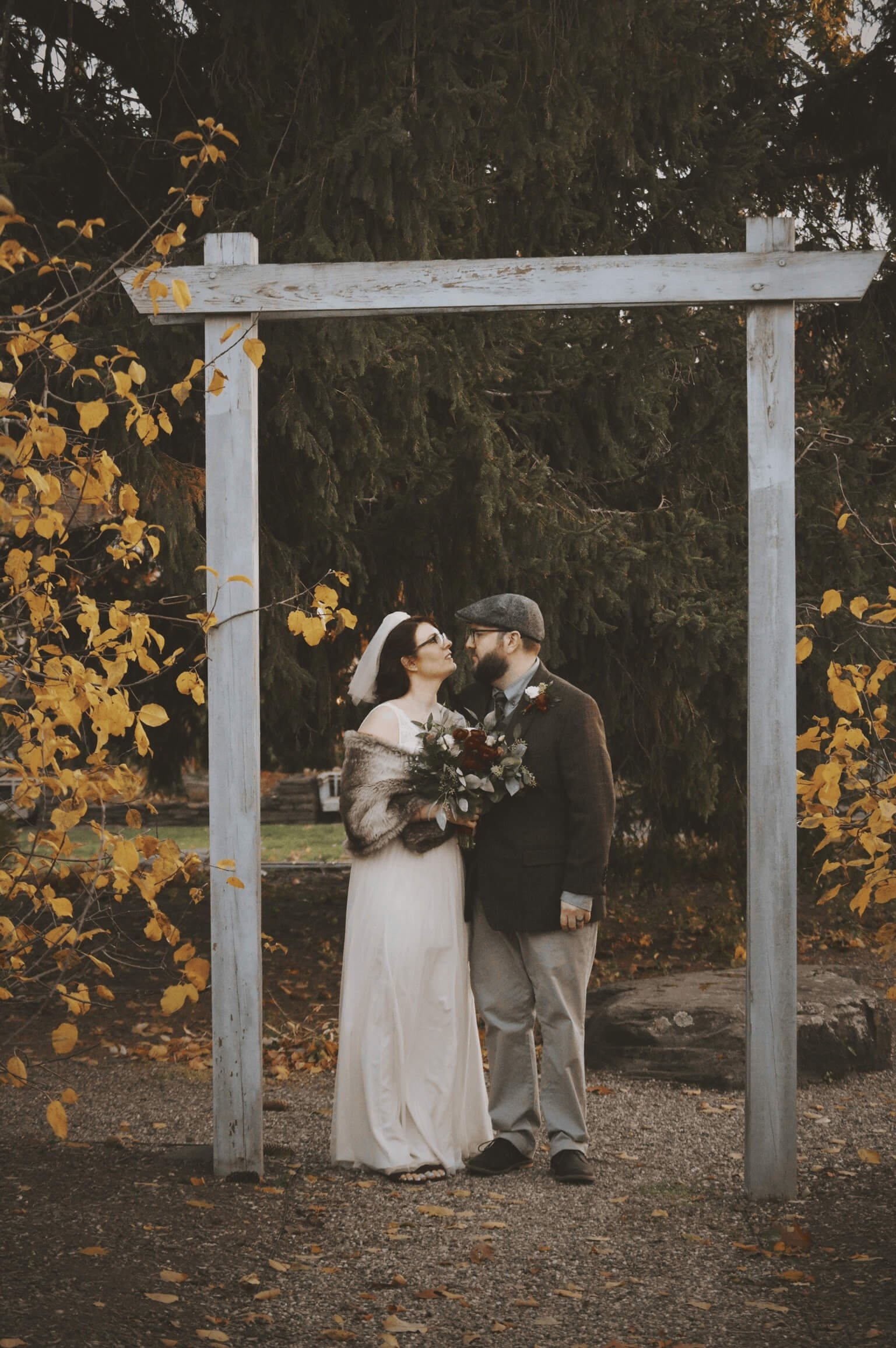 Bride and groom under a rustic wooden arch with golden fall foliage, holding a burgundy and sage bouquet, Milkweed Floral Co
