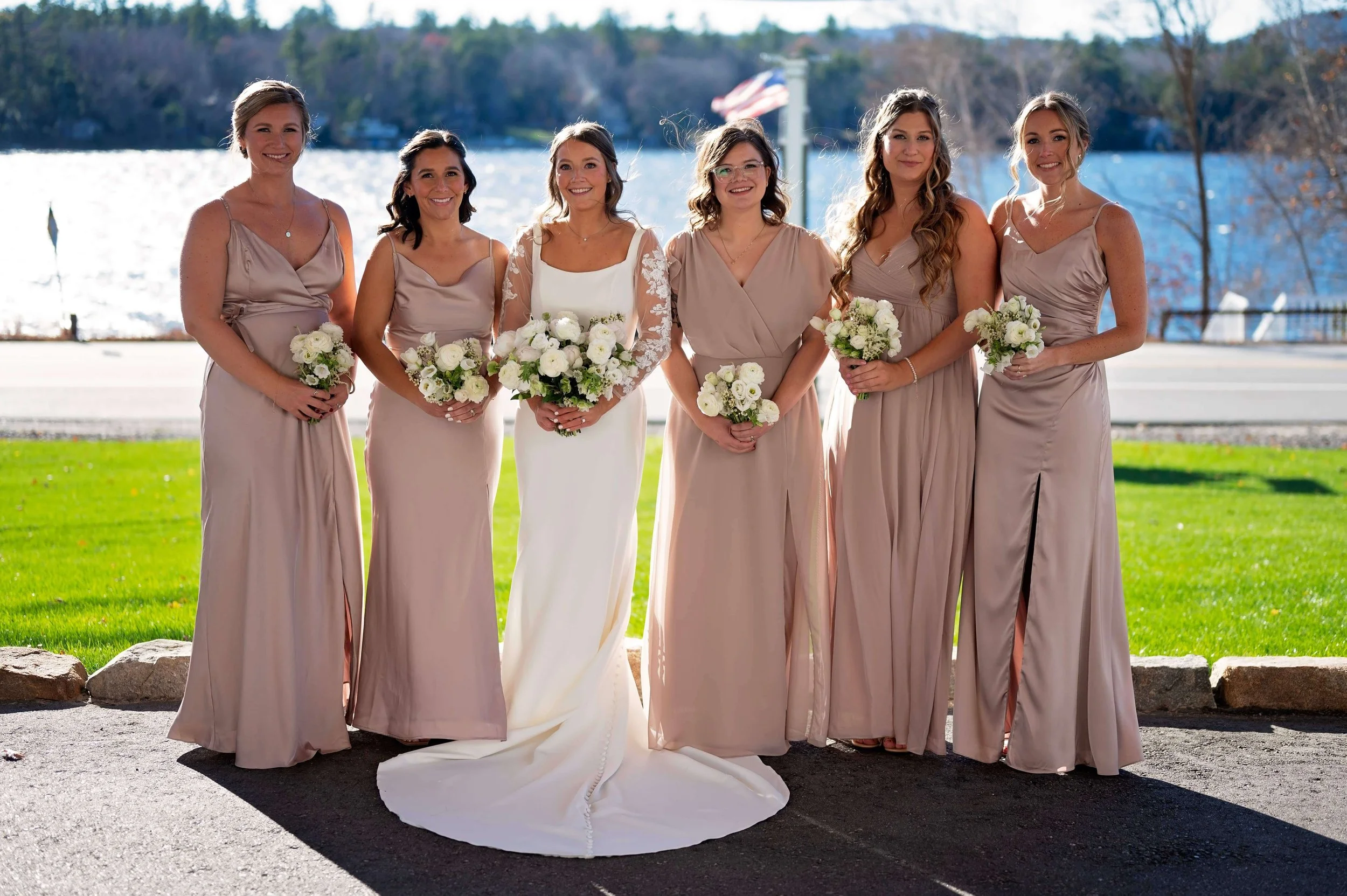 Bride and bridesmaids in taupe gowns holding white ranunculus and garden rose bouquets at Newfound Lake Inn, Bridgewater New Hampshire wedding florals by Milkweed Floral Co