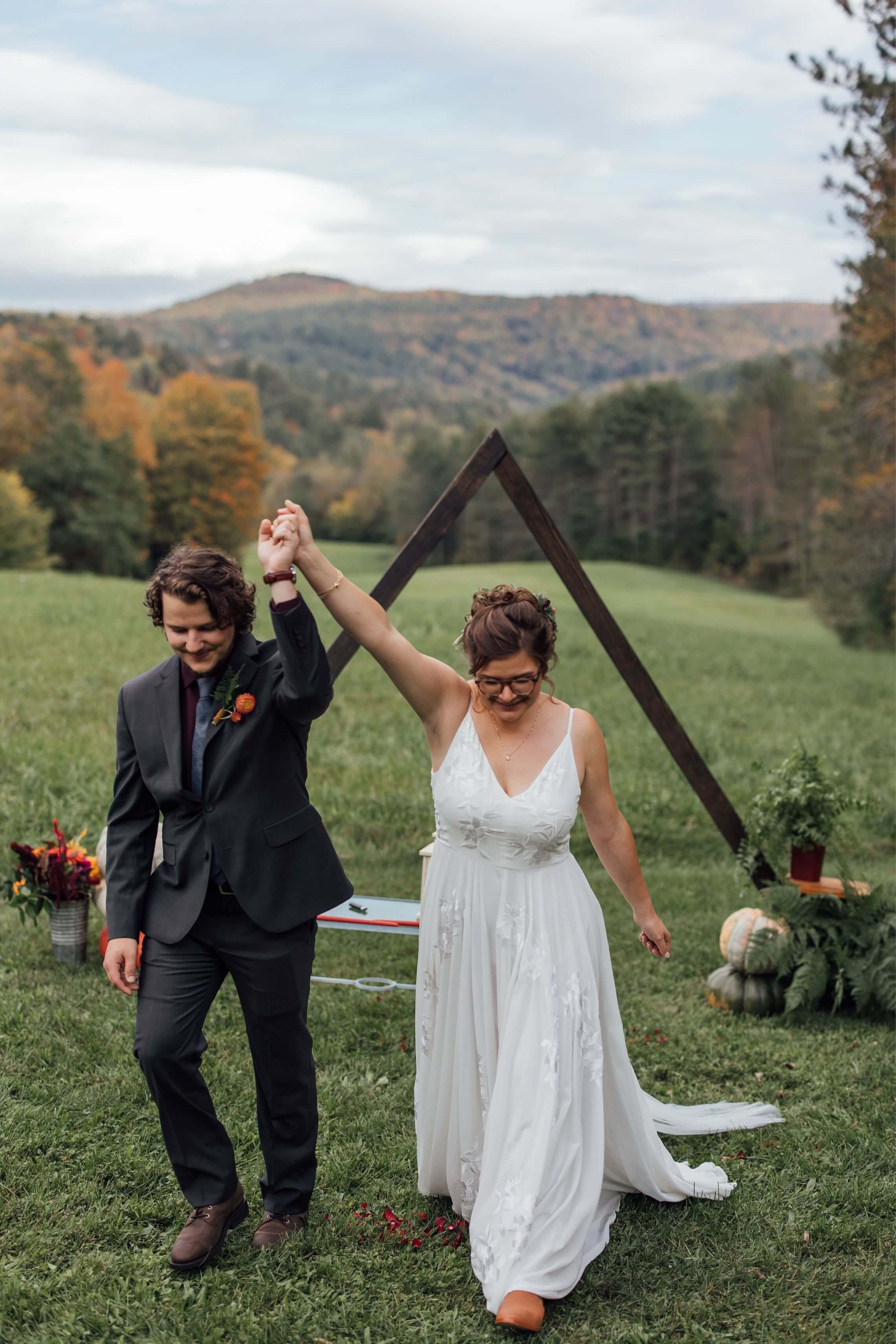Bride and groom celebrating their recessional with hands raised before a triangular wooden arch and fall foliage mountain views at a private Vermont wedding, wedding florals by Milkweed Floral Co