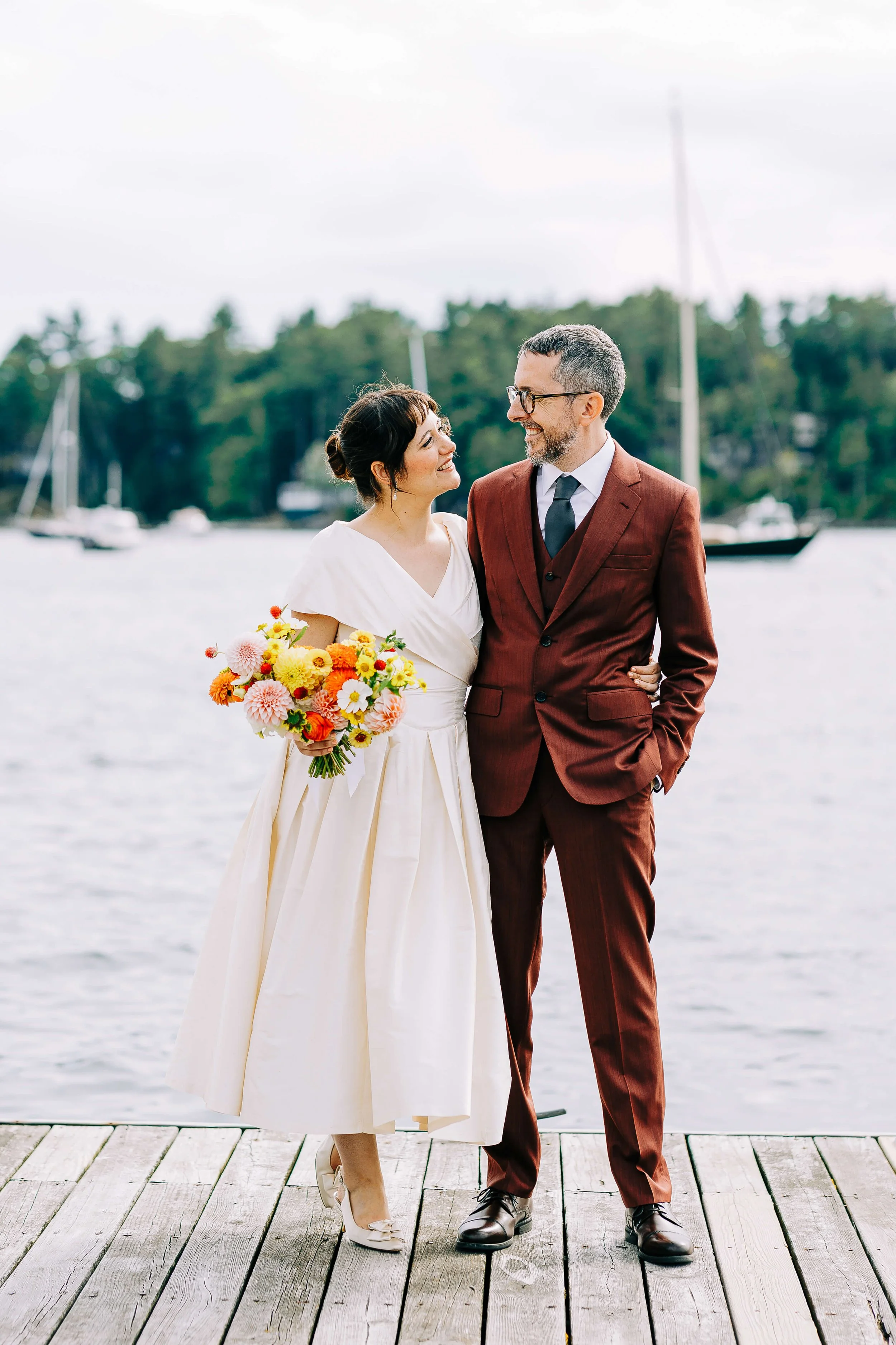 Bride and groom laughing on a harbor dock, bride holding a vibrant orange, yellow, and pink dahlia bouquet, Blue Hill Maine bouquet by Milkweed Floral Co