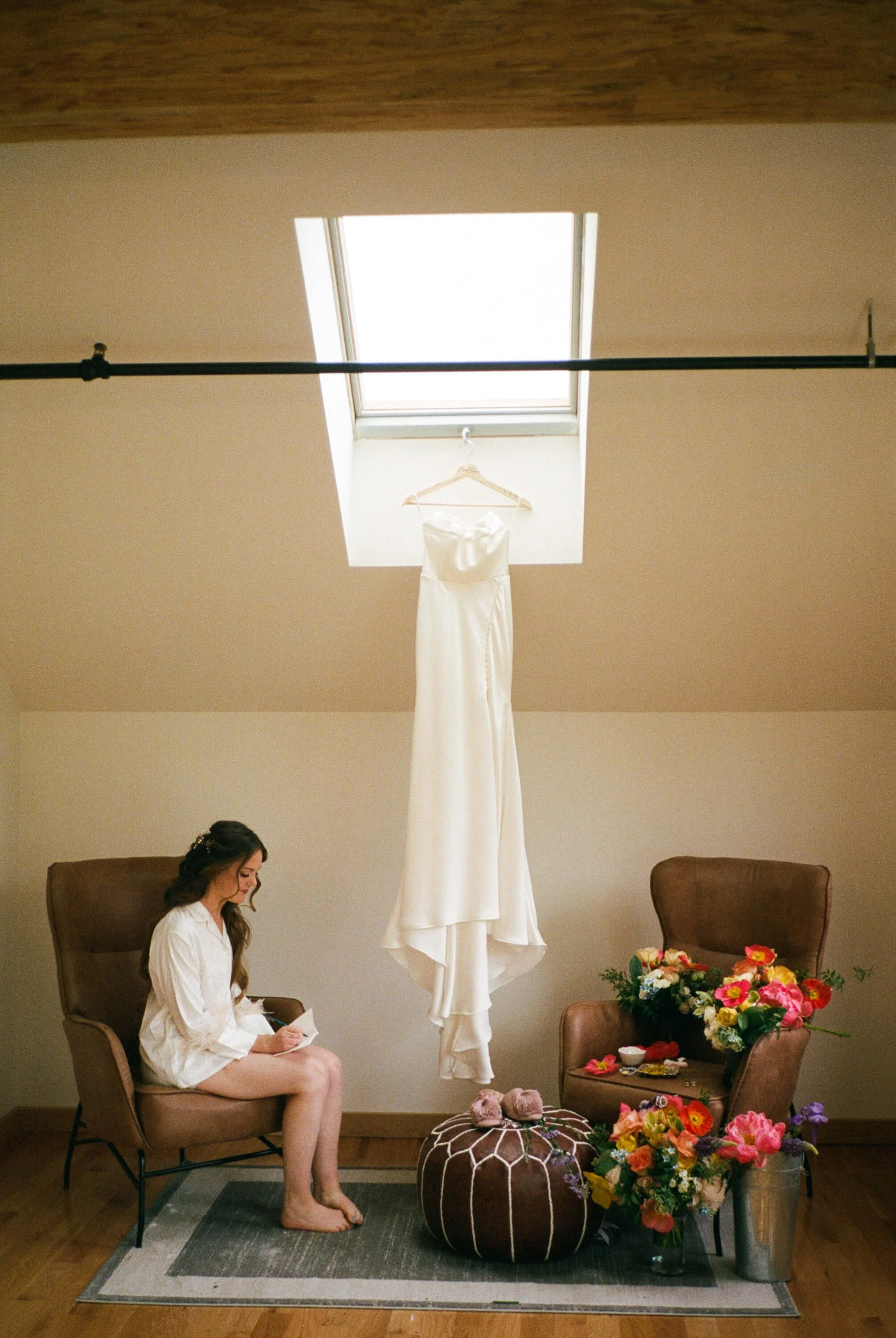 Bride writing vows in a robe beside her hanging wedding dress, with vibrant wildflower arrangements of poppies and tulips nearby, getting ready detail by Milkweed Floral Co