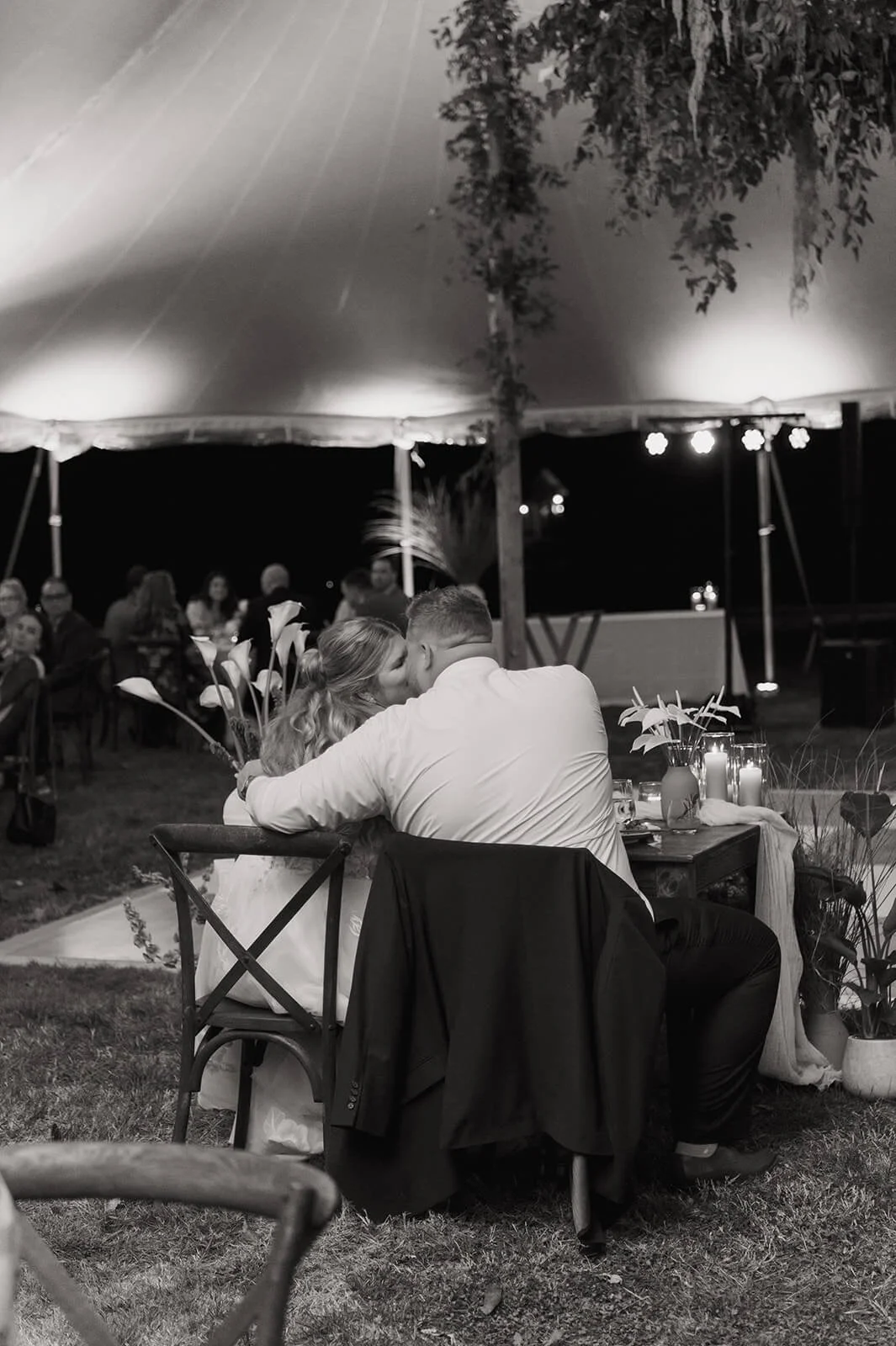 Black and white photo of bride and groom kissing at their sweetheart table with calla lily arrangements and candles under a hanging greenery installation, Monson Massachusetts backyard wedding, Milkweed Floral Co
