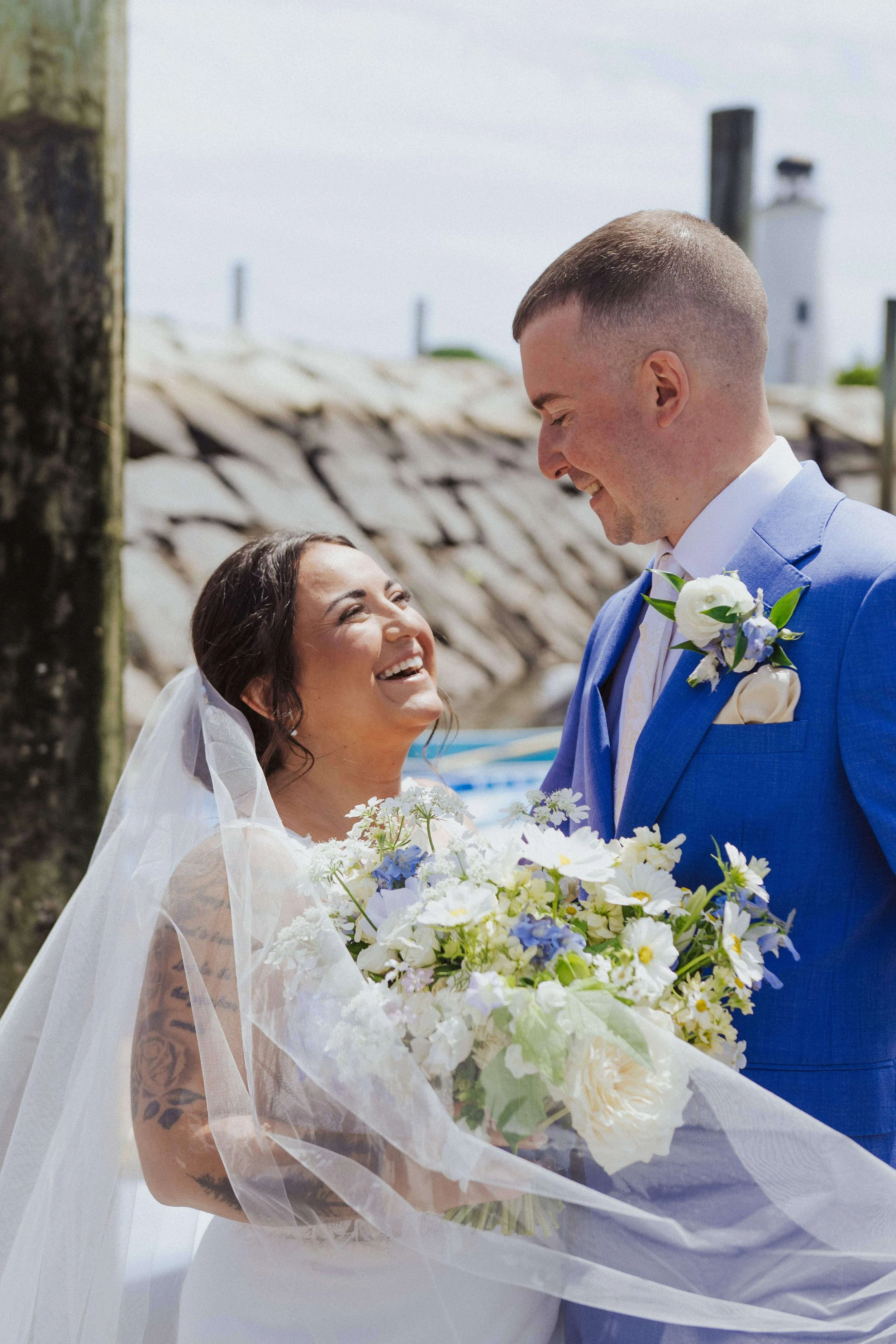 Joyful bride and groom smiling at each other, lush bridal bouquet with white and blue flowers, on dock in Kennebunkport, Maine, Milkweed Floral Co