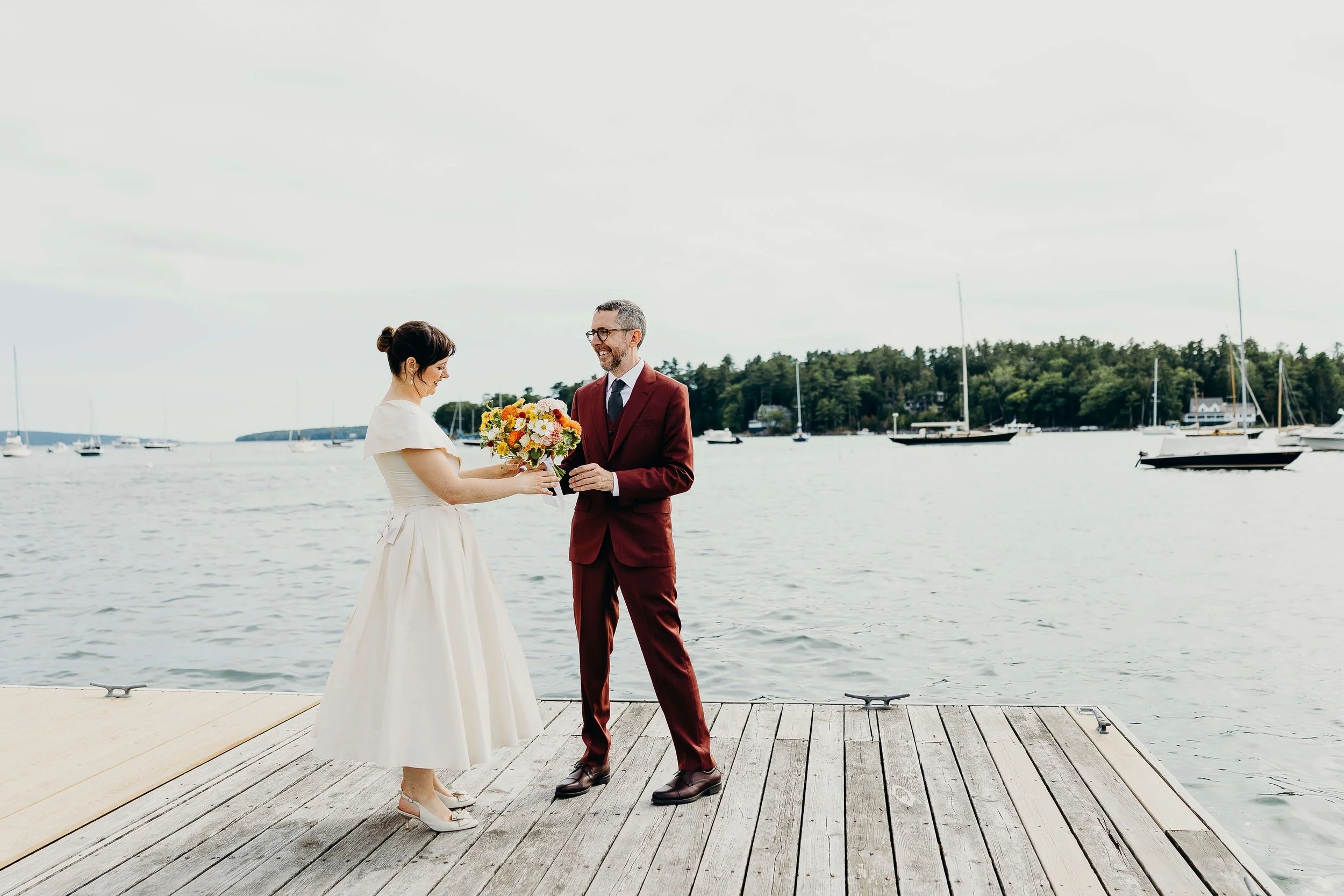 Bride and groom sharing a joyful moment on a coastal Maine dock, bride holding a vibrant orange, yellow, and pink wildflower bouquet, wedding florals by Milkweed Floral Co