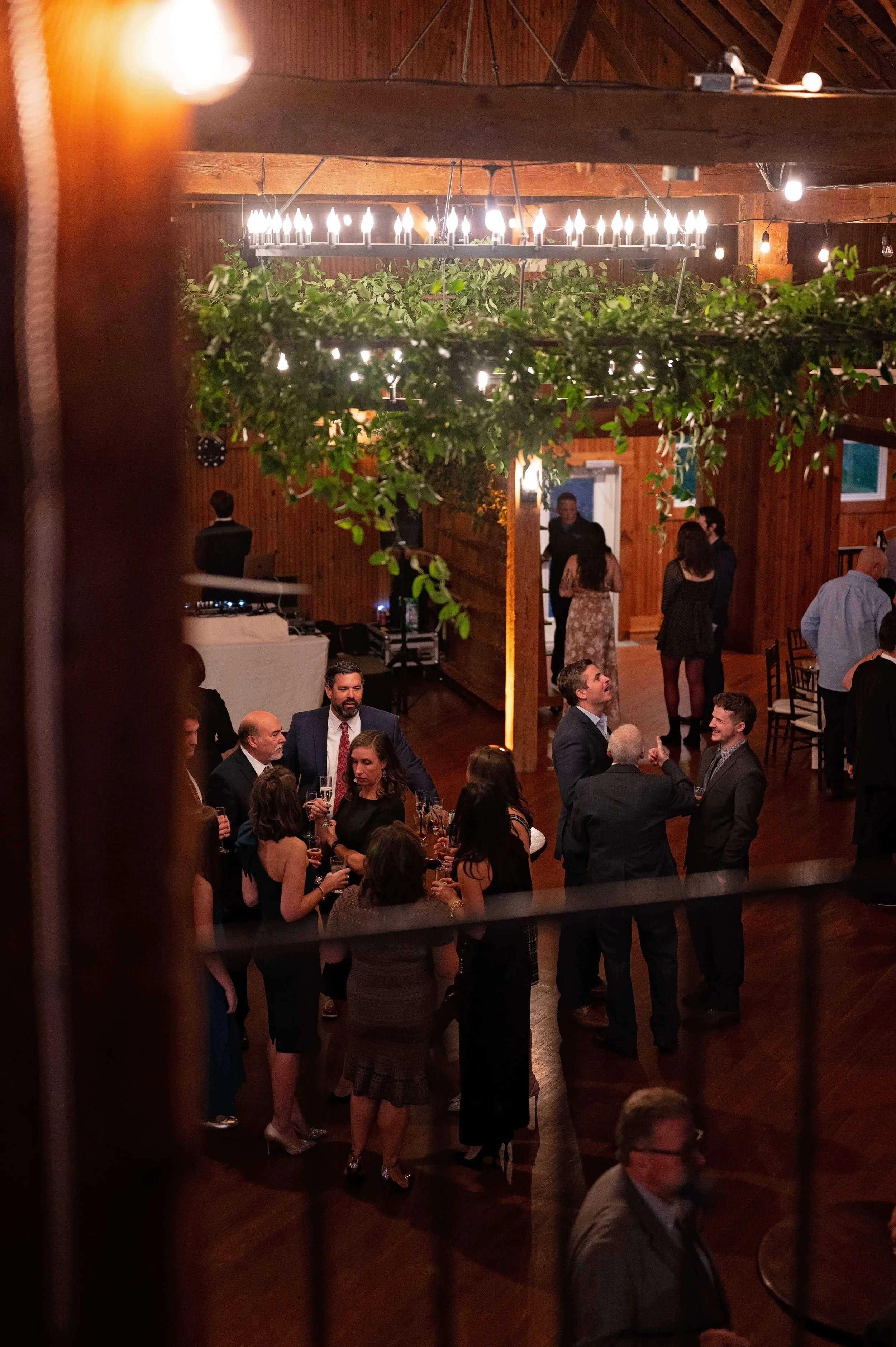 Wedding guests mingling beneath a lush greenery chandelier installation at Newfound Lake Inn, Bridgewater New Hampshire wedding florals by Milkweed Floral Co