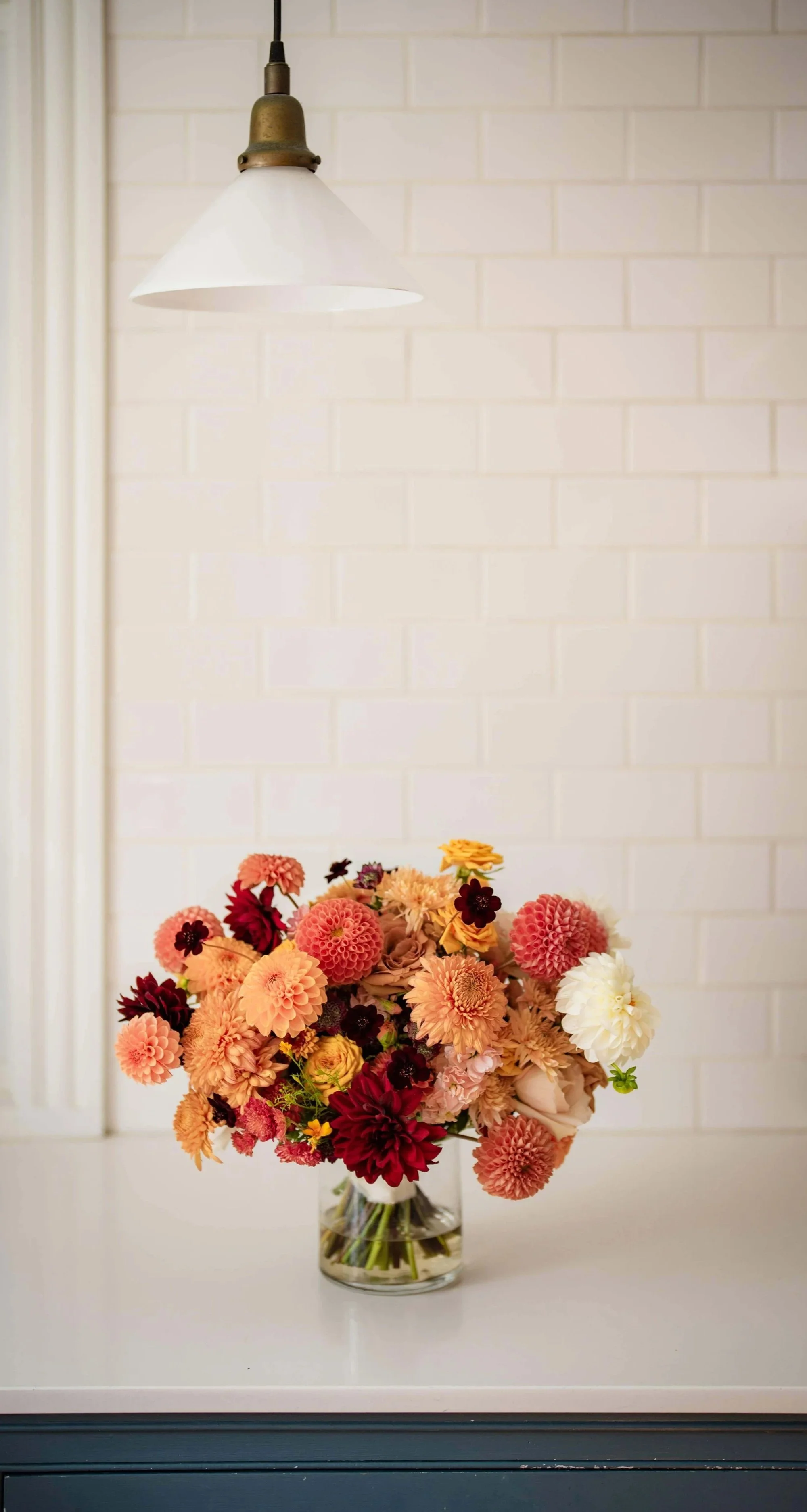 Lush autumn floral arrangement with peach, coral, and burgundy dahlias and ranunculus in a glass vase on a white kitchen counter, Milkweed Floral Co