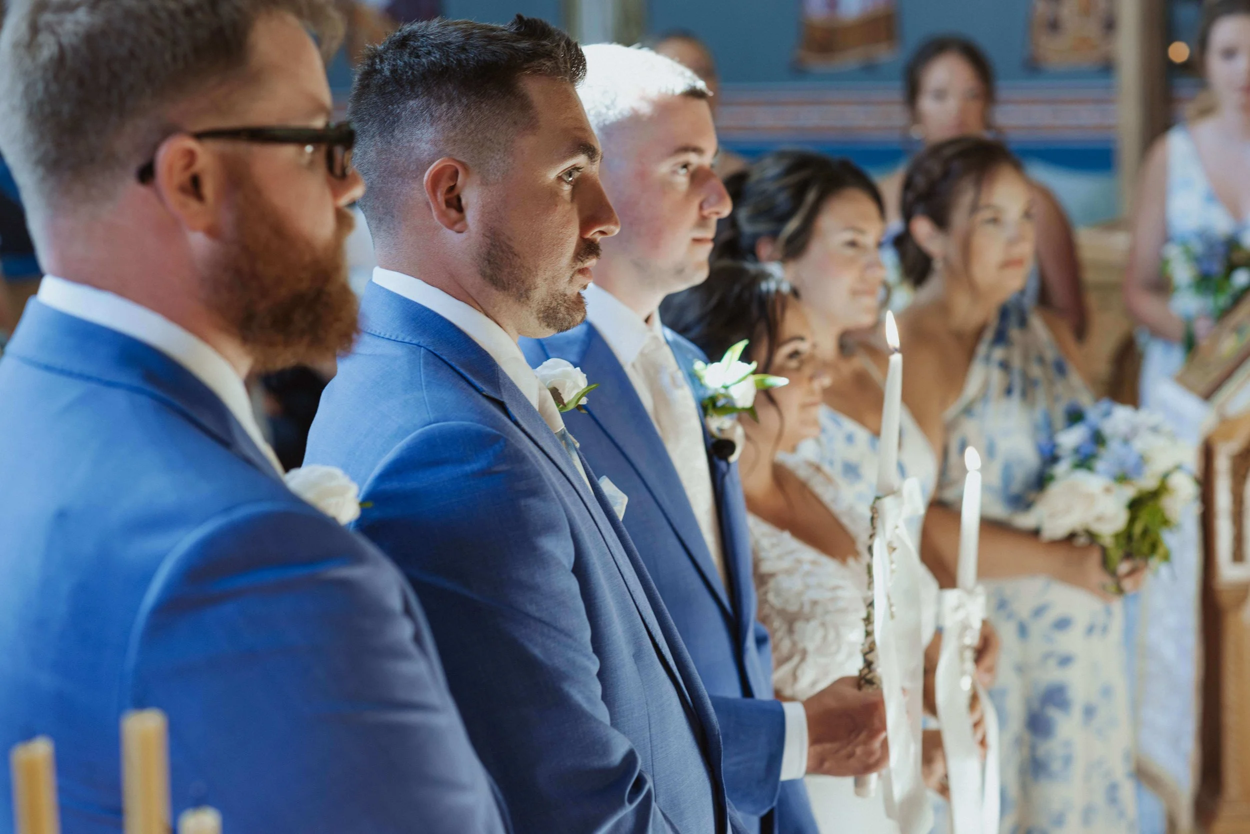 Groomsmen in cobalt blue suits with white ranunculus boutonnieres during the ceremony, bridesmaids in blue floral dresses visible behind them, Kennebunkport Maine, Milkweed Floral Co