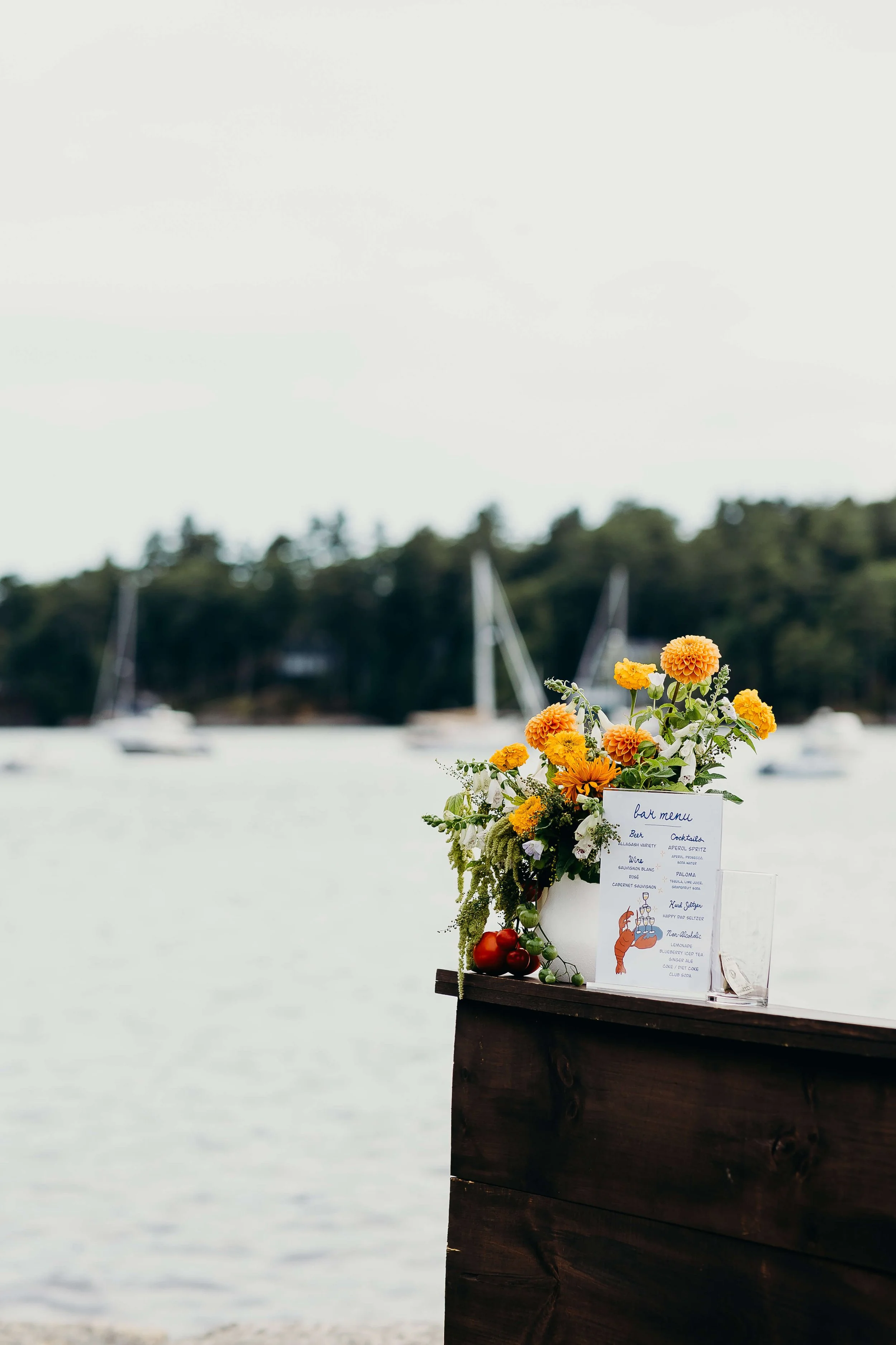 Yellow dahlia and white wildflower bar arrangement overlooking a sailboat-dotted harbor at Kollegewidgwok Yacht Club, Blue Hill Maine wedding florals by Milkweed Floral Co