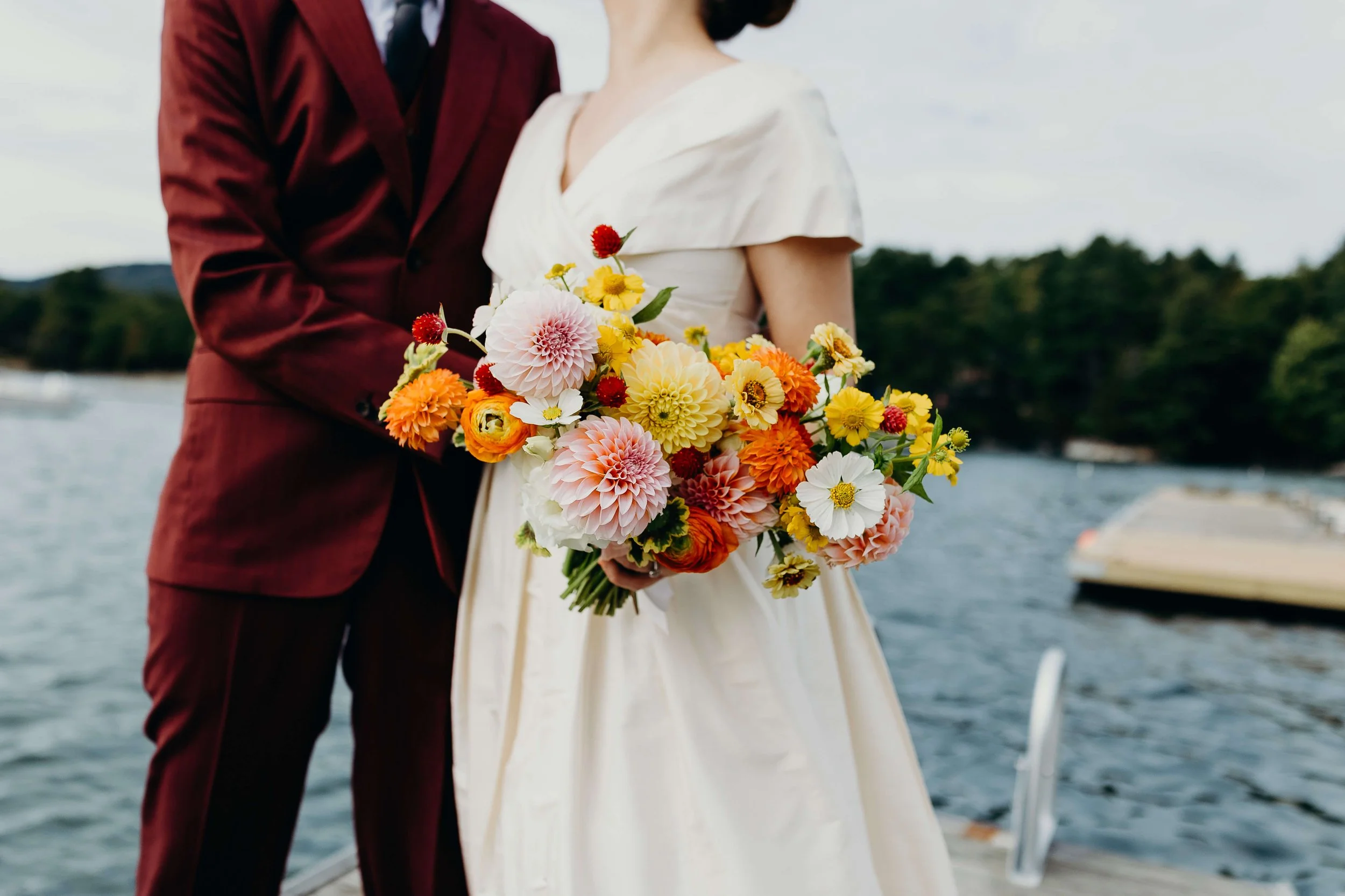 Vibrant wildflower bridal bouquet with pink and orange dahlias, yellow zinnias, ranunculus, and cosmos held on a waterfront dock at Kollegewidgwok Yacht Club, Blue Hill Maine, Milkweed Floral Co