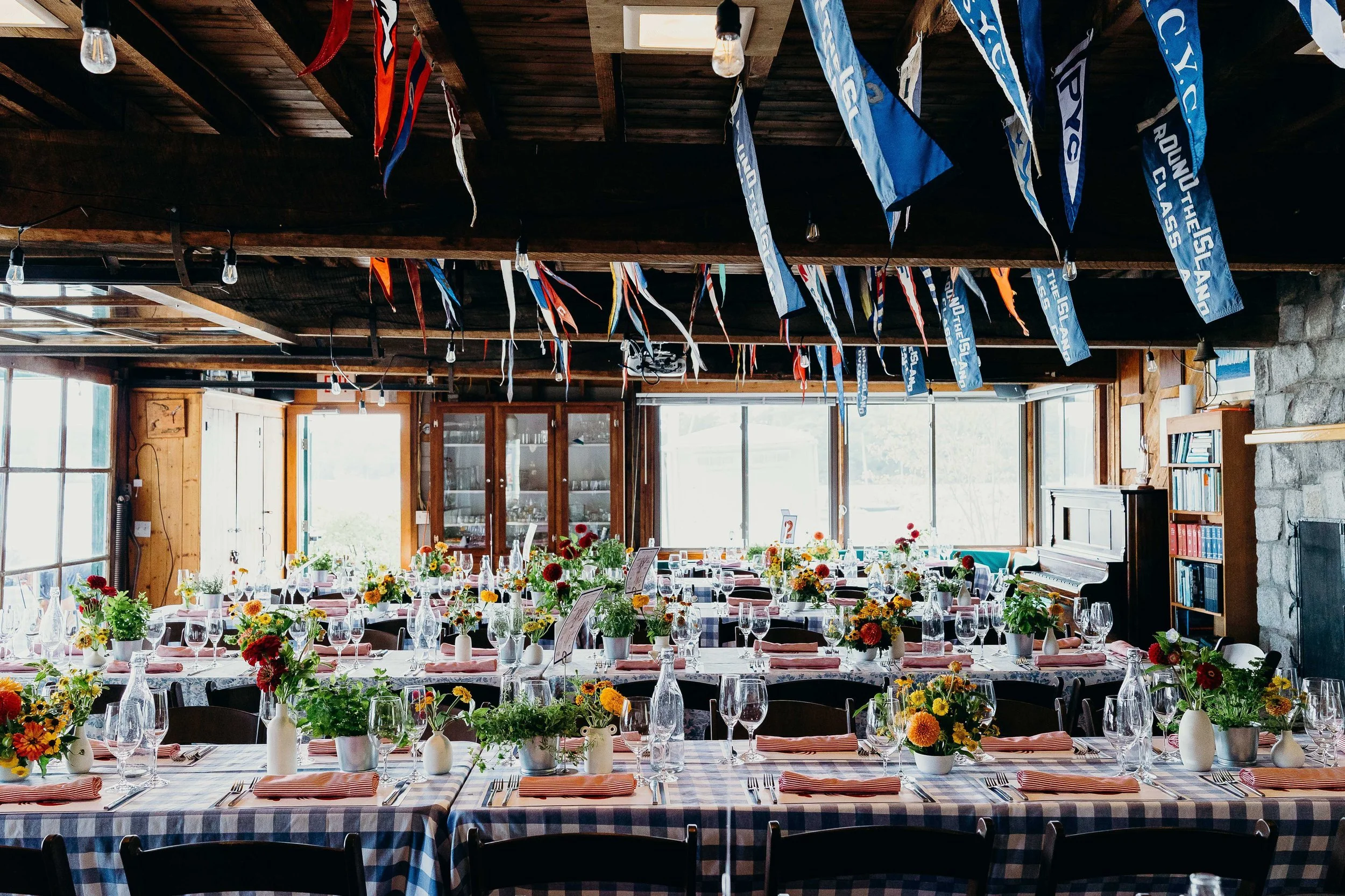 Wedding reception room at Kollegewidgwok Yacht Club with long tables set with colorful wildflower and dahlia centerpieces, blue gingham tablecloths, striped napkins, and sailing flags overhead, Blue Hill Maine wedding by Milkweed Floral Co