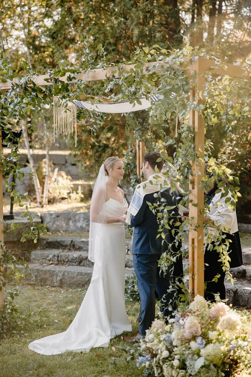 Bride and groom exchanging vows under a lush greenery-draped chuppah with hydrangea and delphinium aisle florals at an outdoor coastal Maine wedding, Milkweed Floral Co