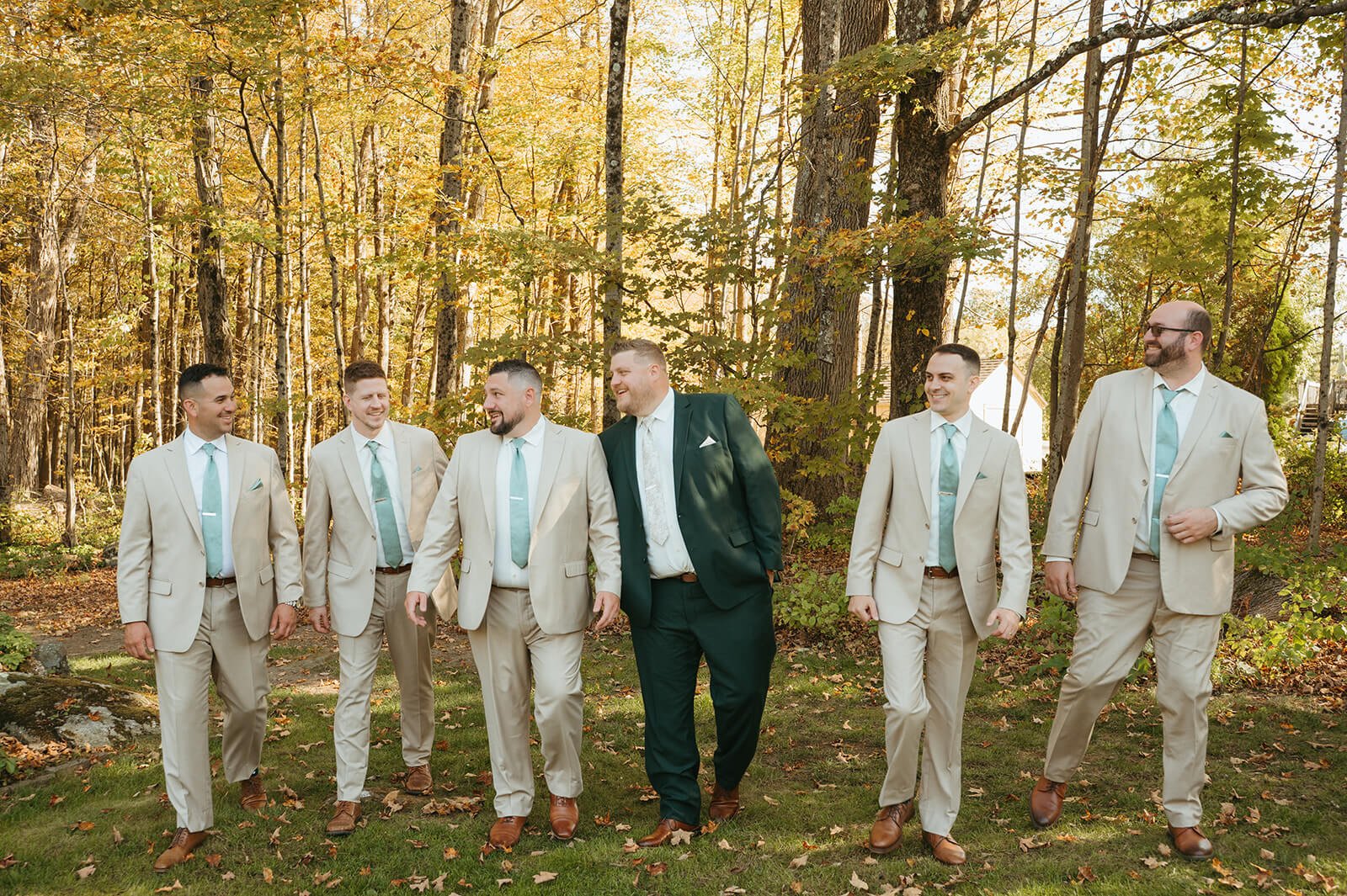 Groom in a forest green suit walking with groomsmen in tan suits and sage ties against a backdrop of golden fall foliage, Monson Massachusetts backyard wedding by Milkweed Floral Co