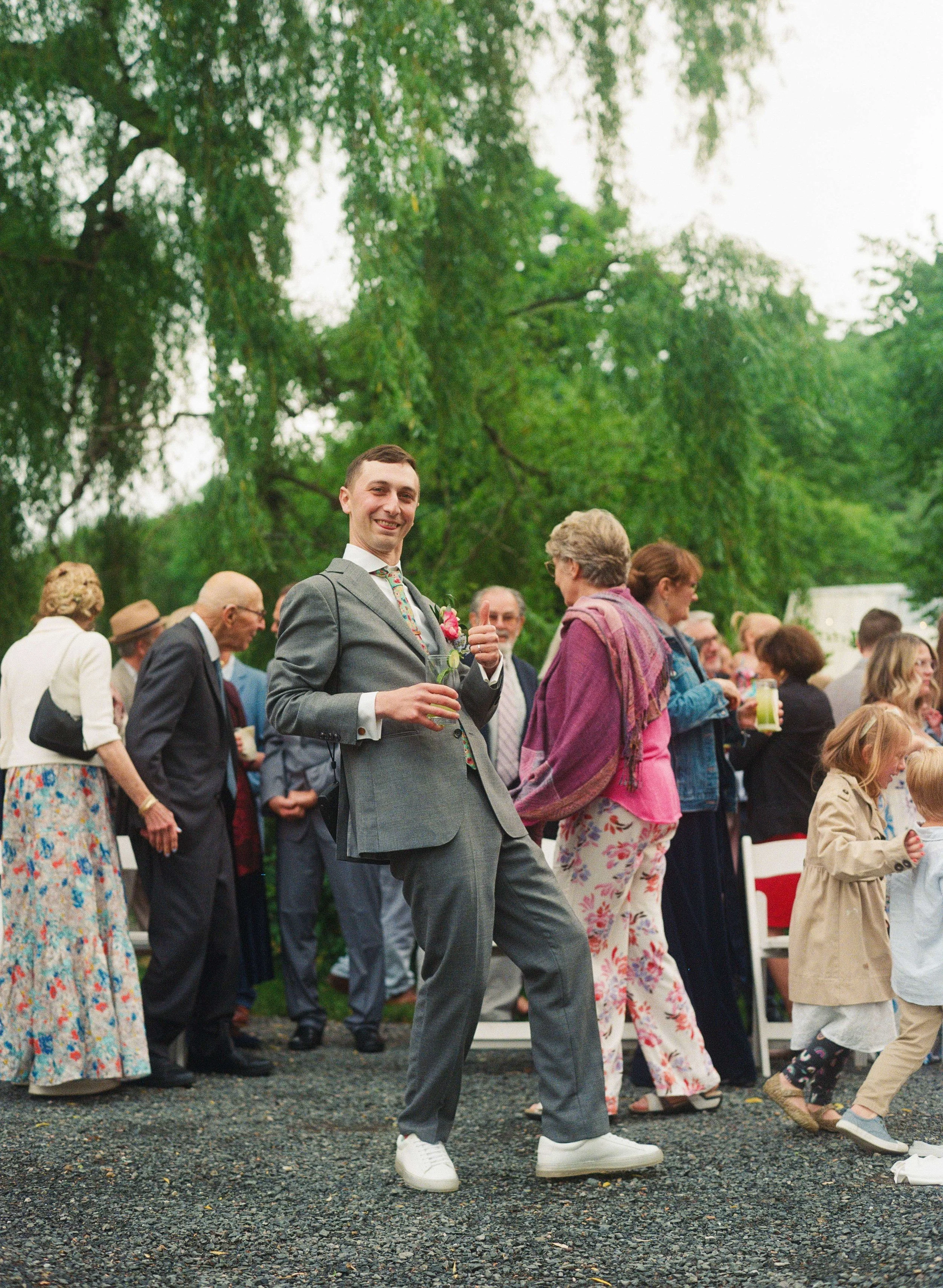 Smiling groom in a grey suit with a colorful wildflower boutonniere mingling with guests at an outdoor cocktail hour under a weeping willow, New England wedding by Milkweed Floral Co