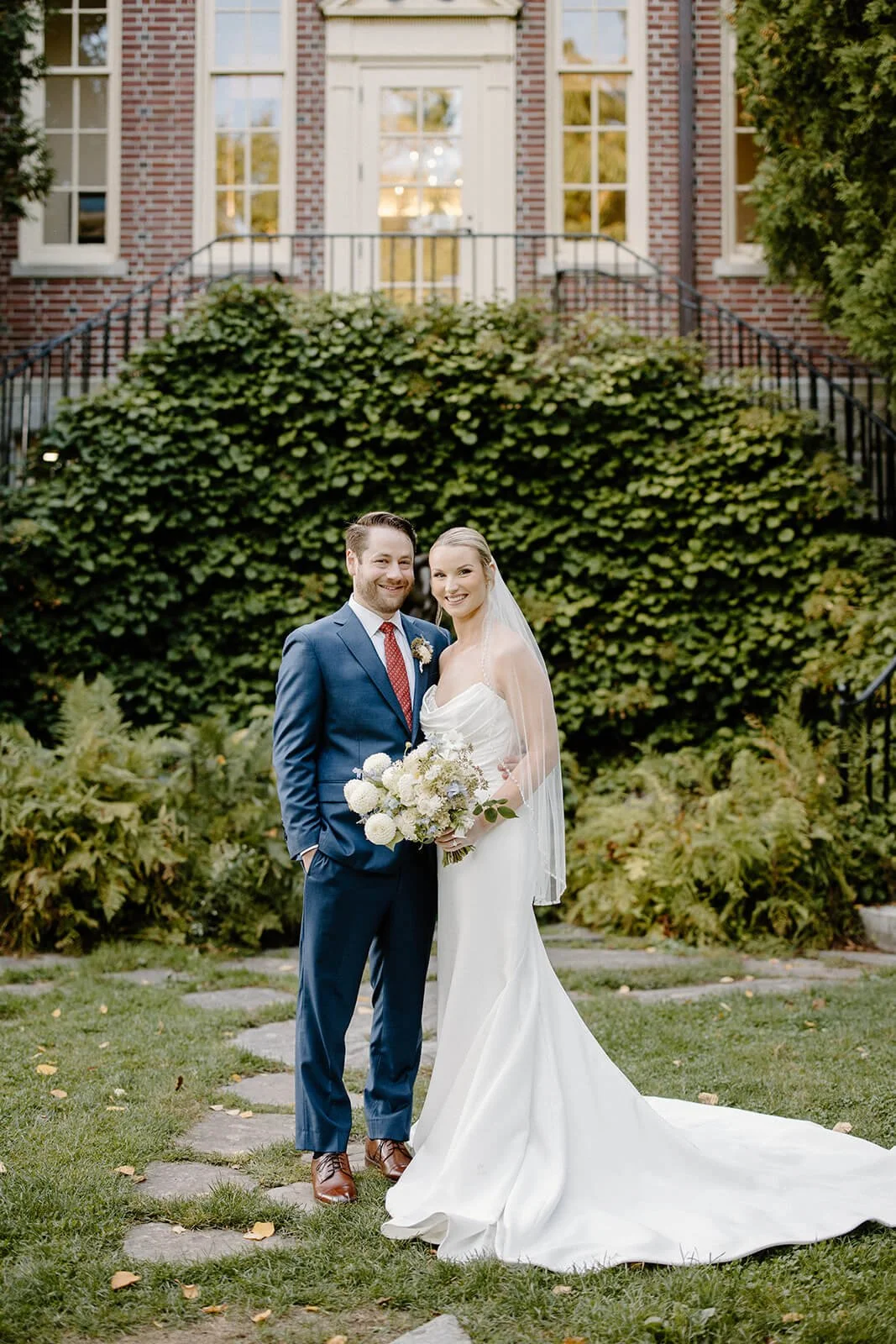Bride and groom portraits in front of Camden Public Library, bride holding a white dahlia and blue delphinium bouquet, Maine wedding by Milkweed Floral Co