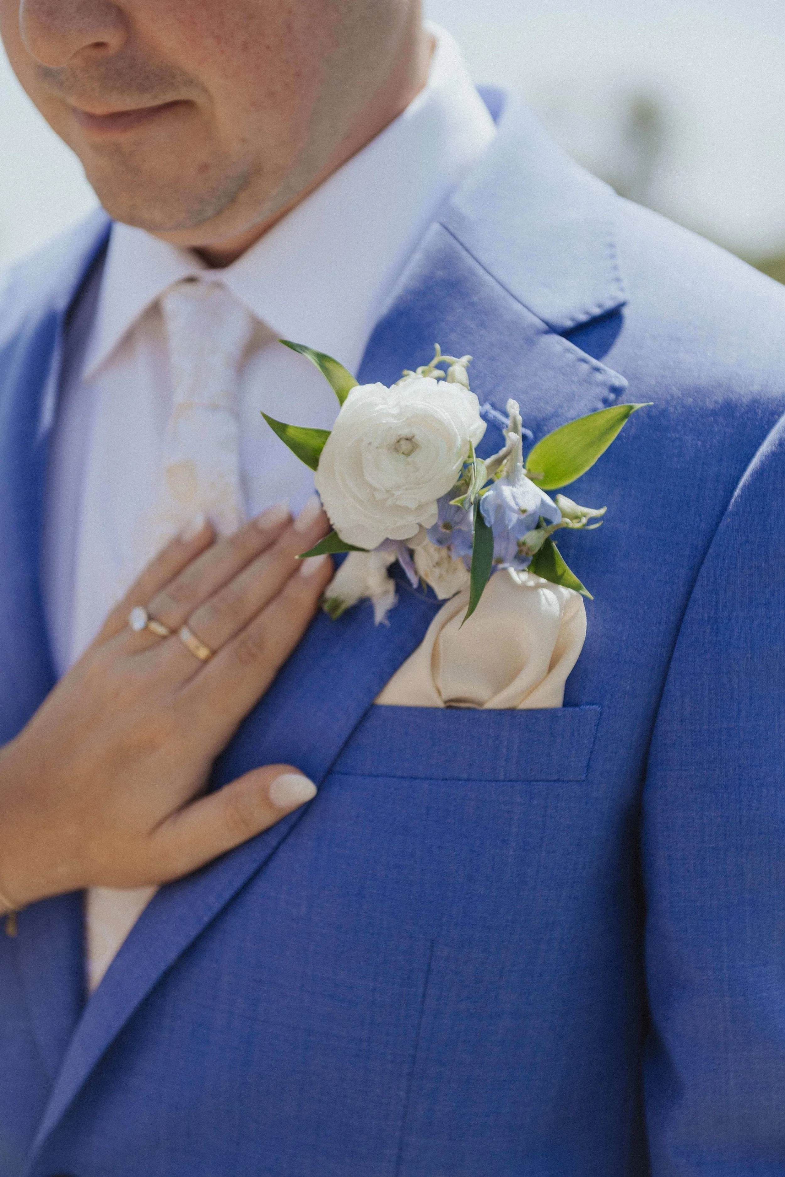 Coastal boutonniere with white ranunculus and blue delphinium on a cobalt blue linen suit, bride's hand resting on groom's chest, Kennebunkport Maine wedding by Milkweed Floral Co