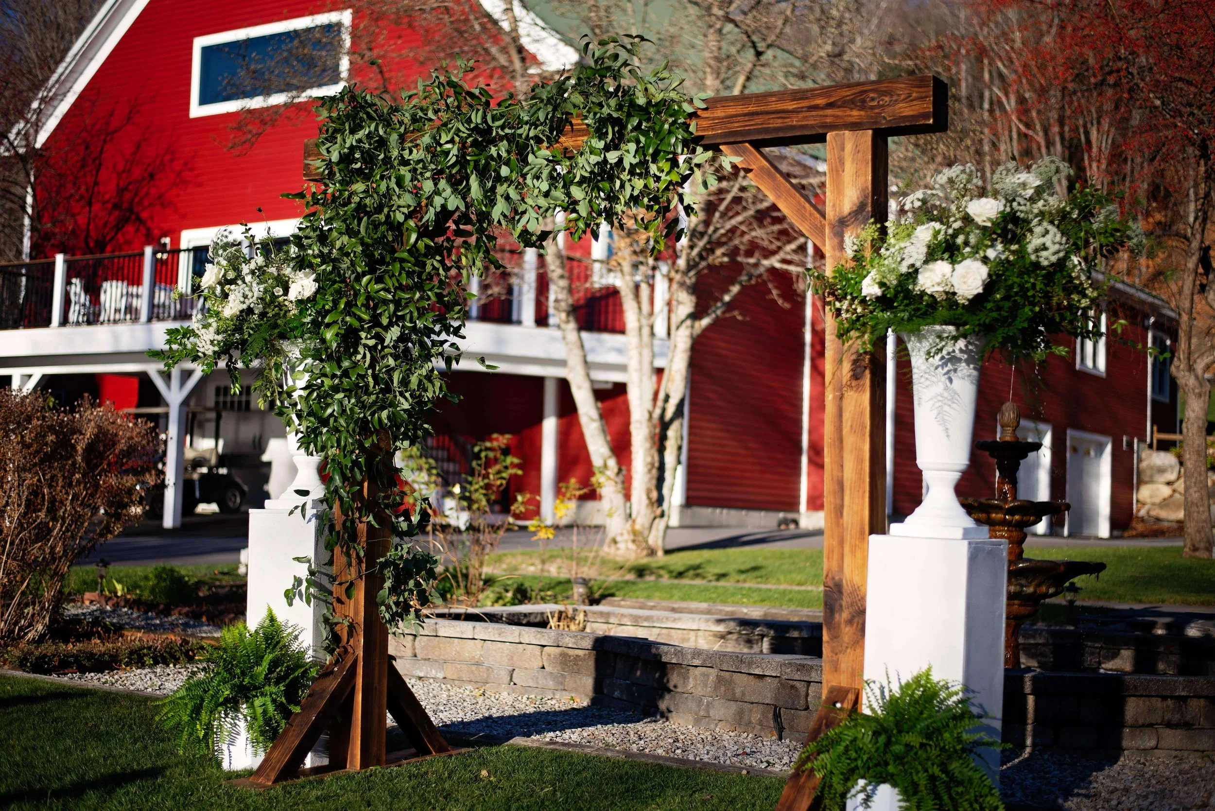 Rustic wooden wedding ceremony arbor with cascading eucalyptus and greenery, flanked by white urn arrangements, red barn backdrop at Newfound Lake Inn, Bridgewater New Hampshire, Milkweed Floral Co